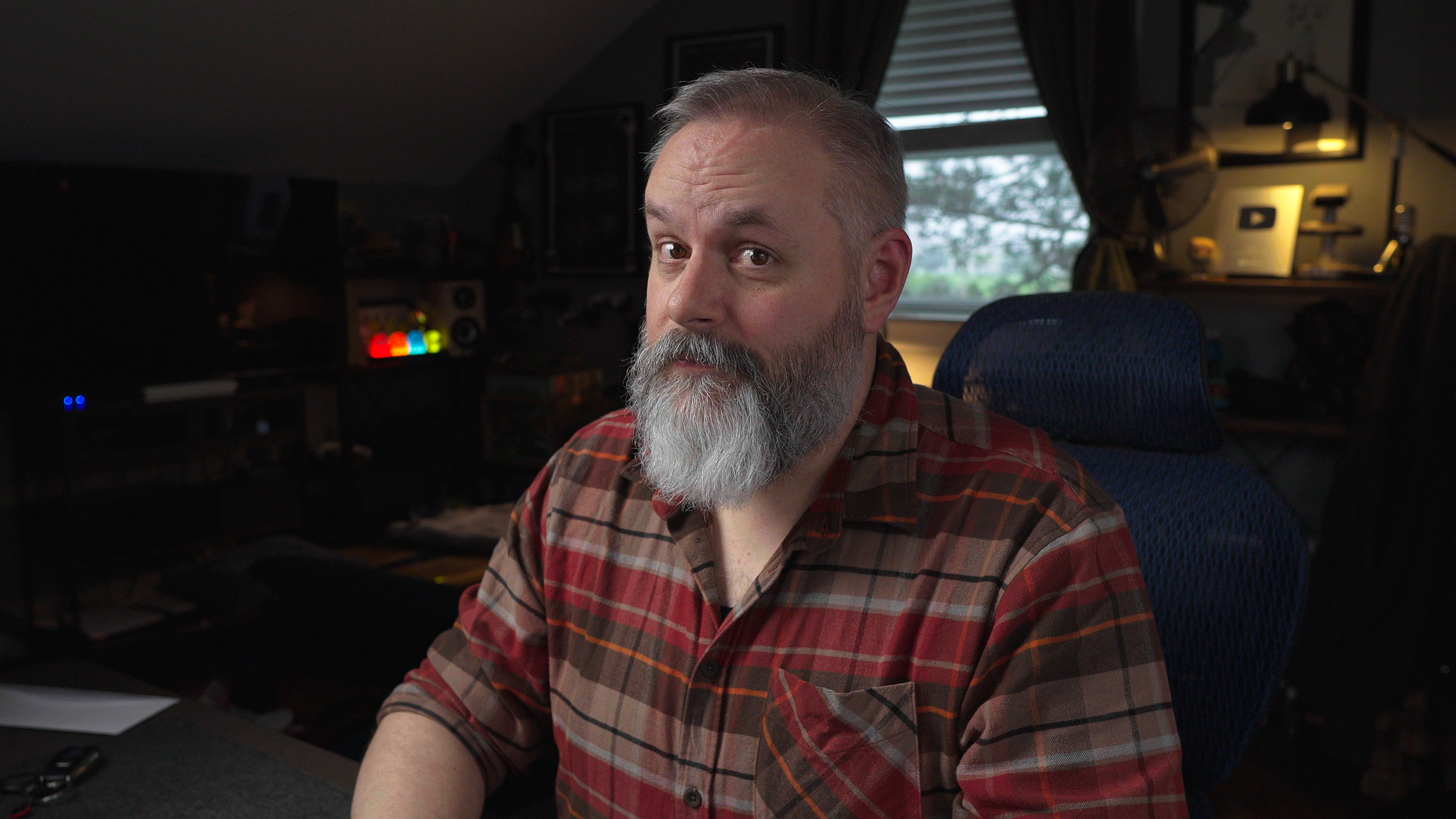 A man with gray hair and a beard, wearing a red and brown plaid shirt, sitting in a dimly lit room next to a window with a view of trees outside. The room has electronic equipment and monitors in the background.