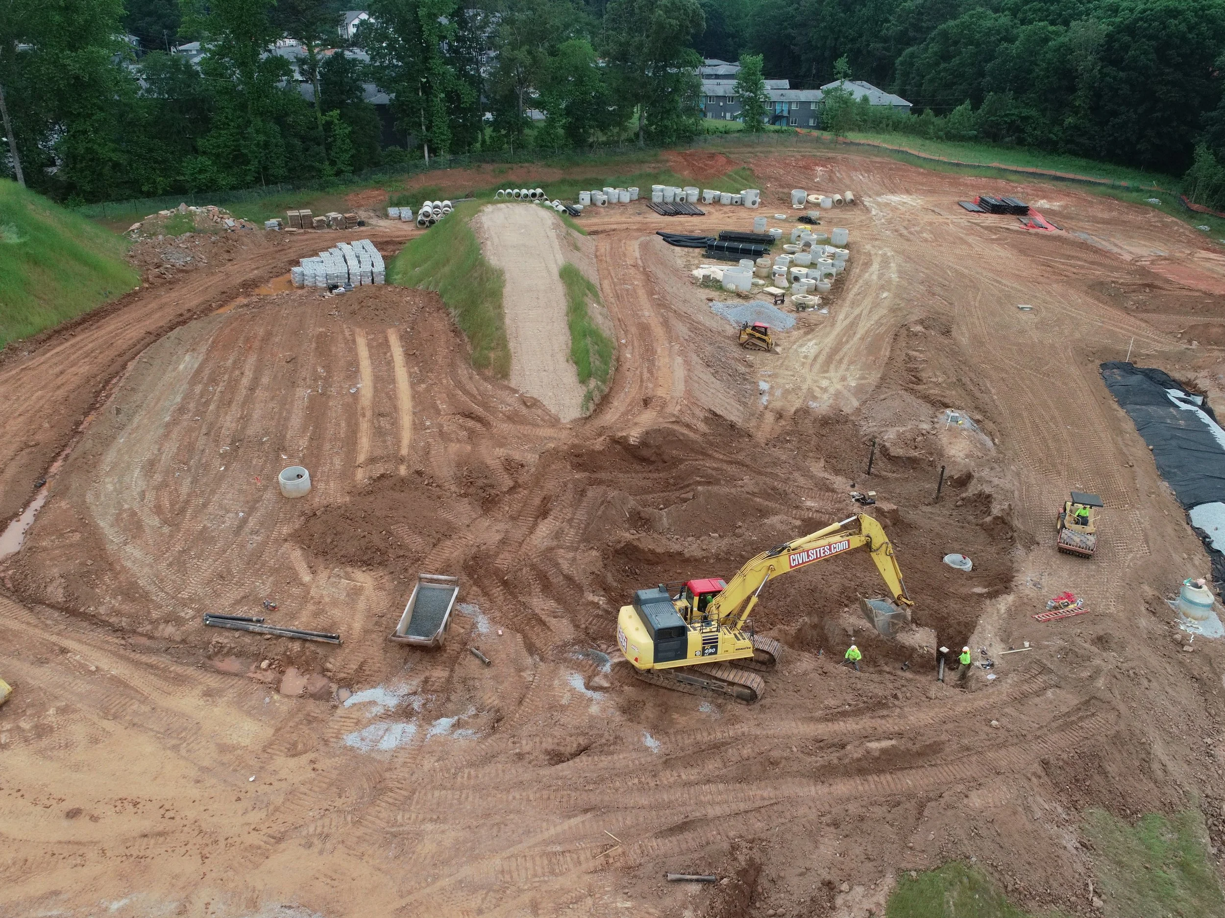Construction site with dirt, machinery, and materials, surrounded by trees and residential buildings in the background.