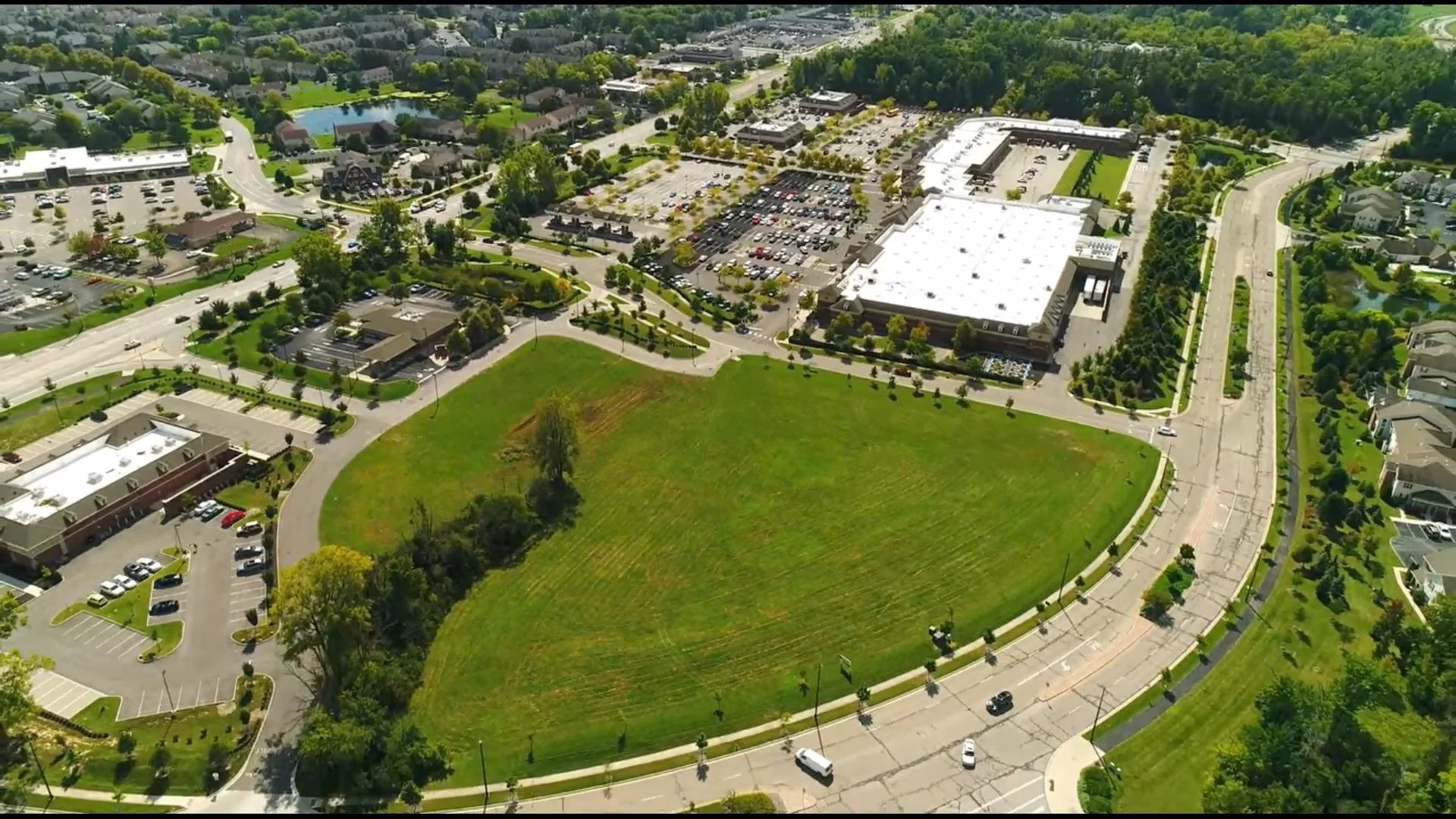 Aerial view of a shopping mall complex surrounded by parking lots, green areas, and residential neighborhoods, with a wooded area in the background.