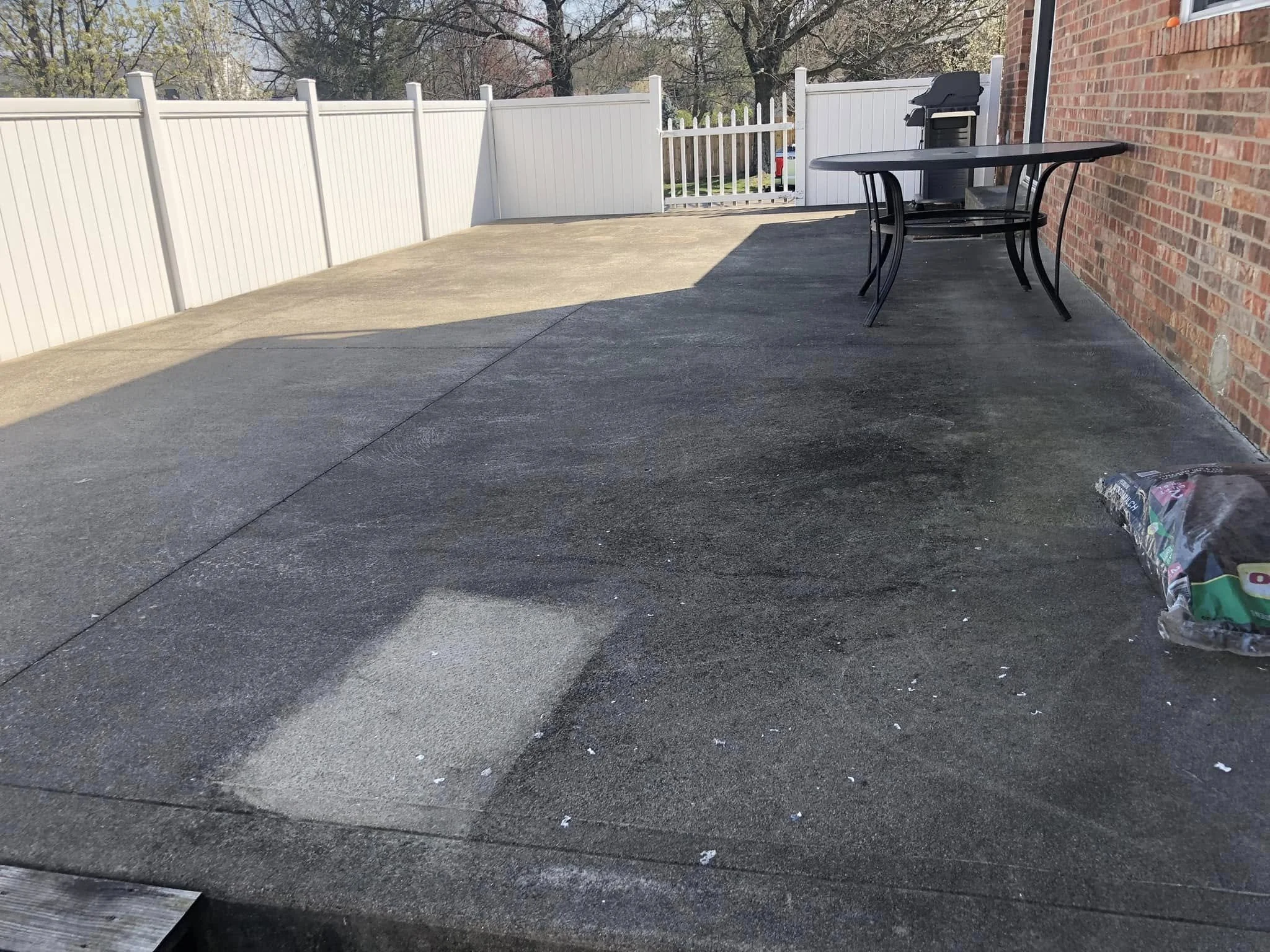 The image shows a backyard patio with a concrete floor, a white fence surrounding the yard, and a brick wall on the right side. There is a black outdoor table and a grill in the background, with a dog food and water dish on the ground in the lower right corner.