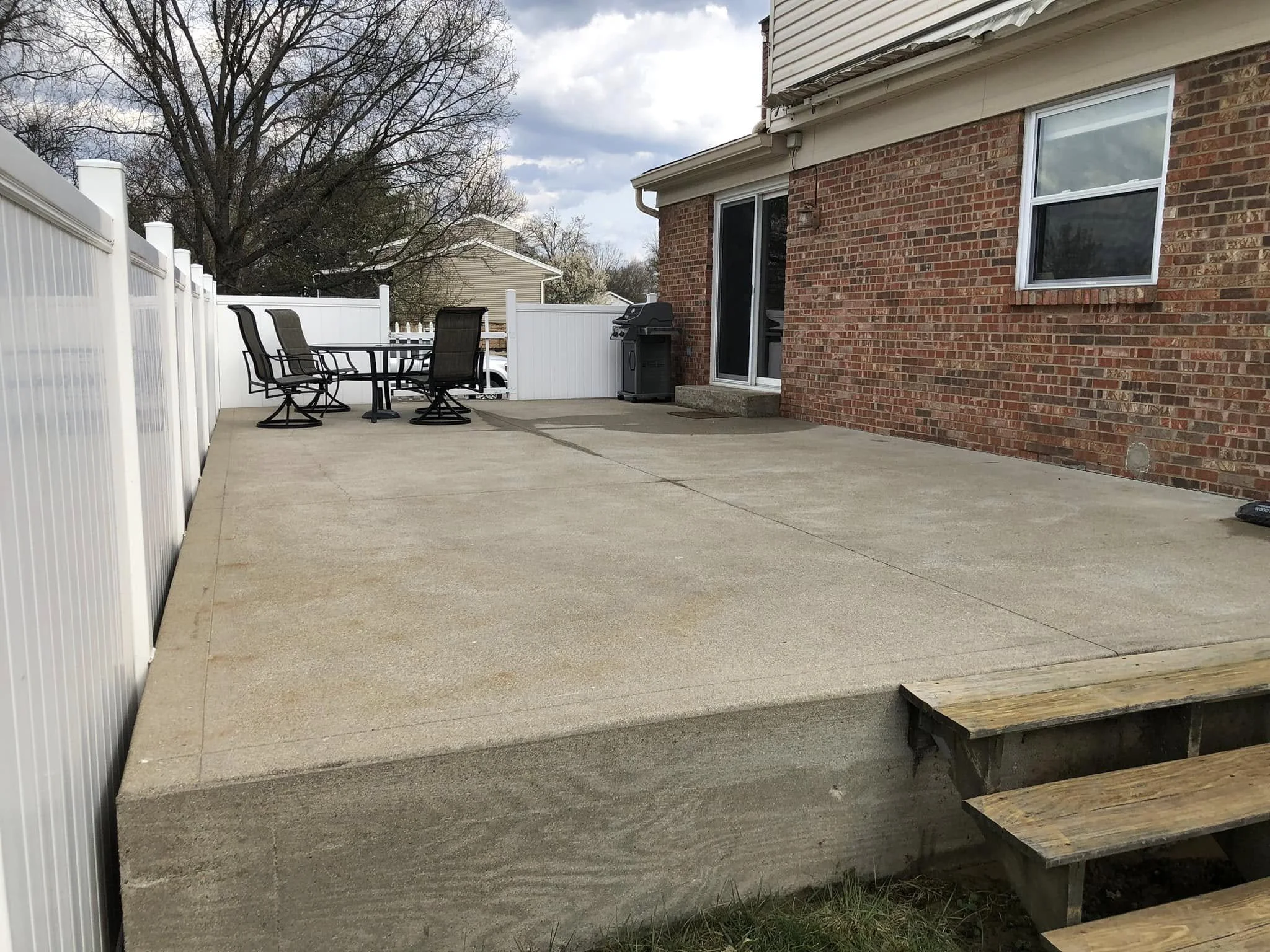 Backyard patio with concrete surface, outdoor table with black metal chairs, a barbecue grill, and a white privacy fence. The house wall is made of red bricks with a window and sliding glass door. There are trees and a cloudy sky in the background.