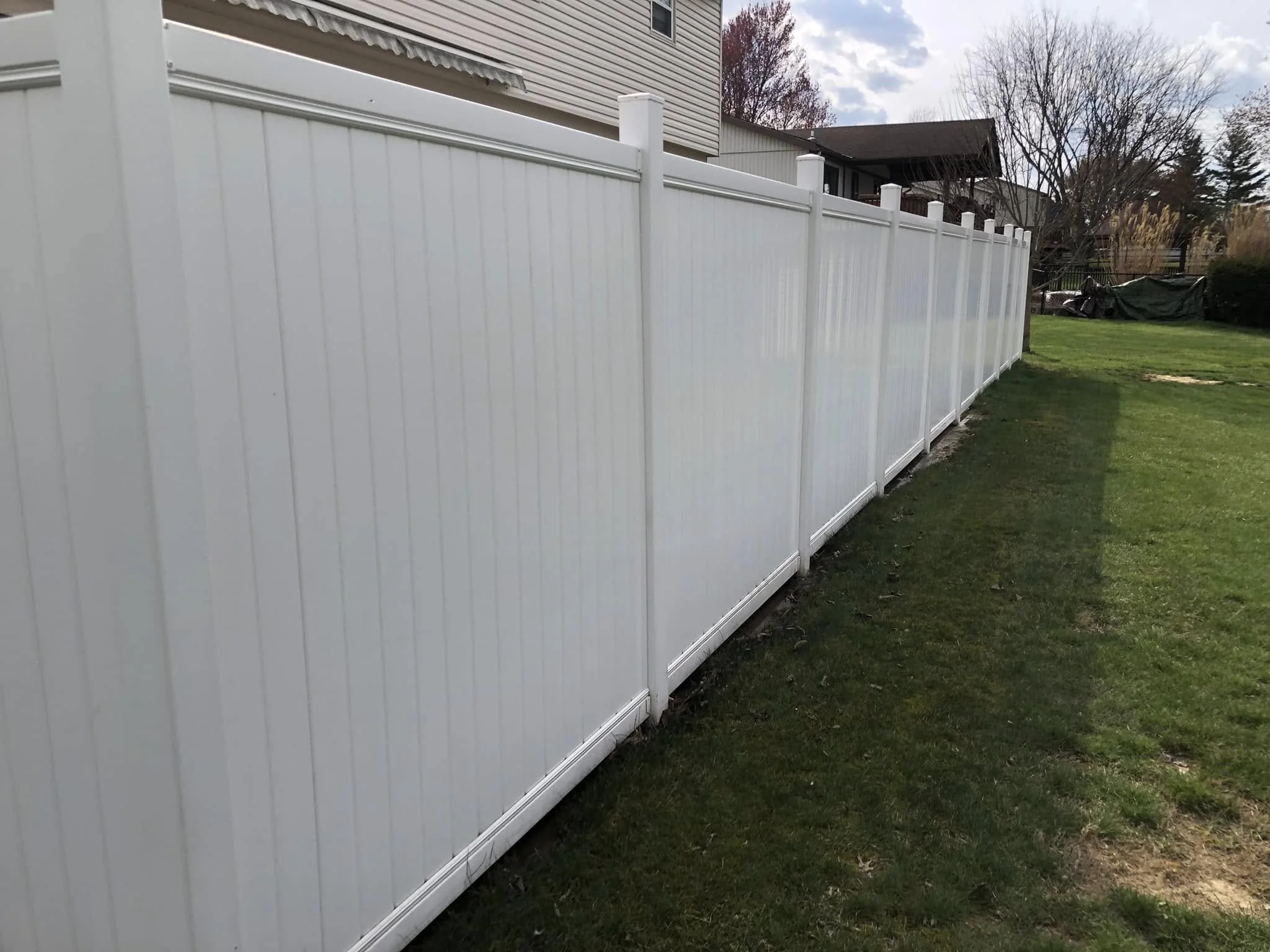 White vinyl privacy fence in a backyard with a house and trees in the background, under a partly cloudy sky.