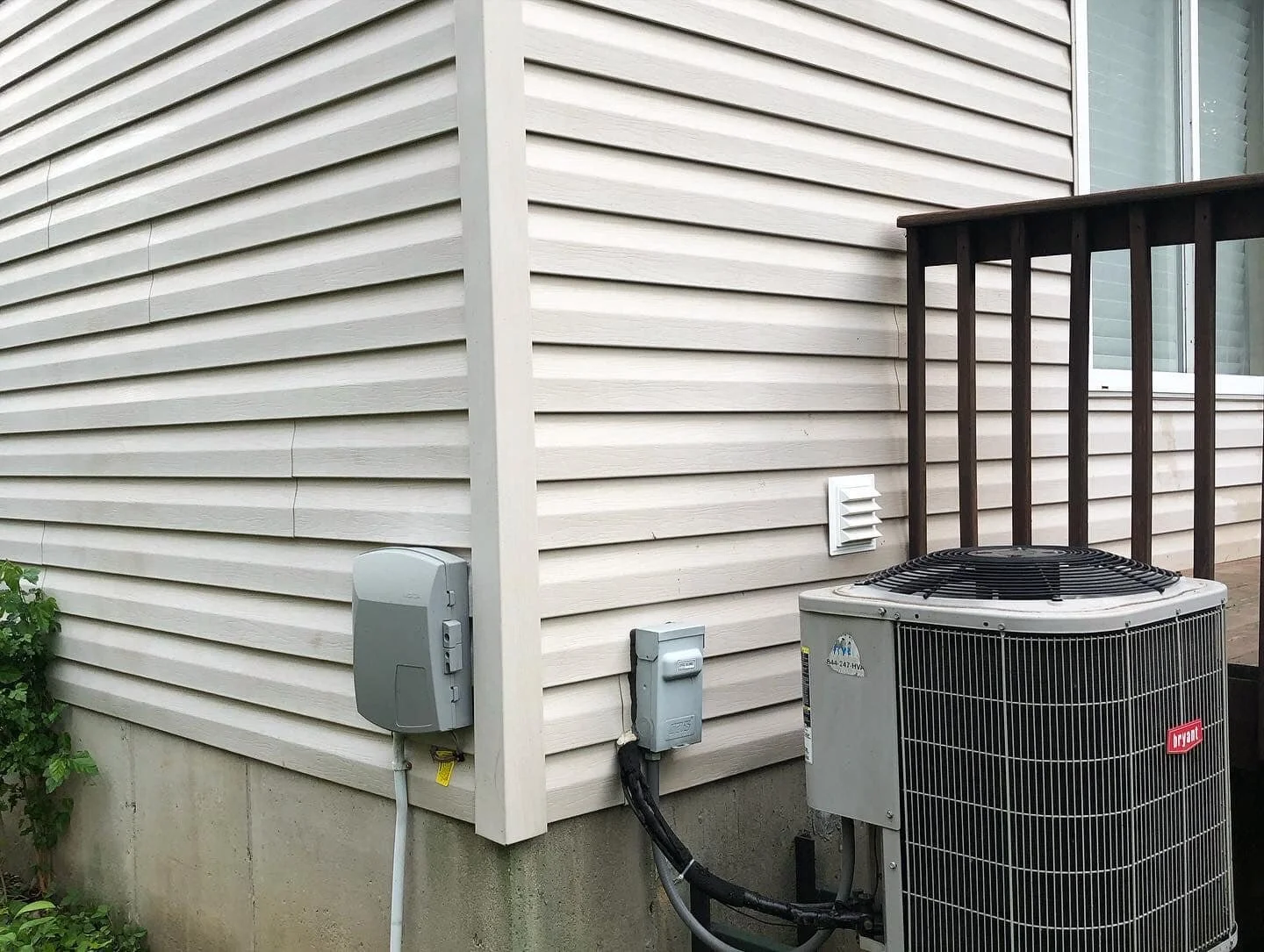 Close-up of the side of a house with beige vinyl siding, an HVAC unit, electrical boxes, and a small vent, with a wooden deck railing nearby.
