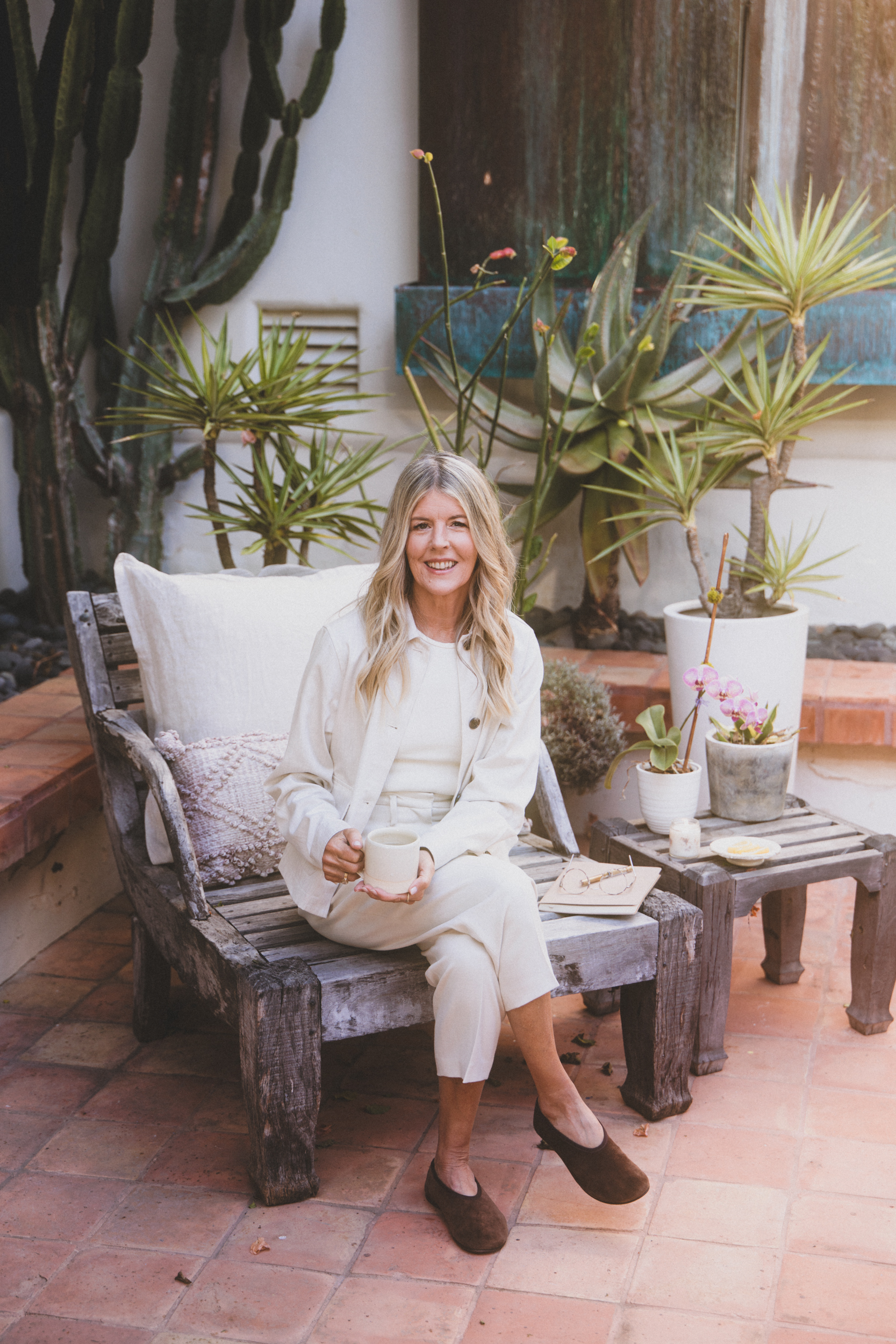 A woman with long blonde hair sitting on a rustic wooden bench in an outdoor patio area with terracotta tiles, surrounded by large potted plants, including succulents and cacti, holding a white mug and smiling at the camera.