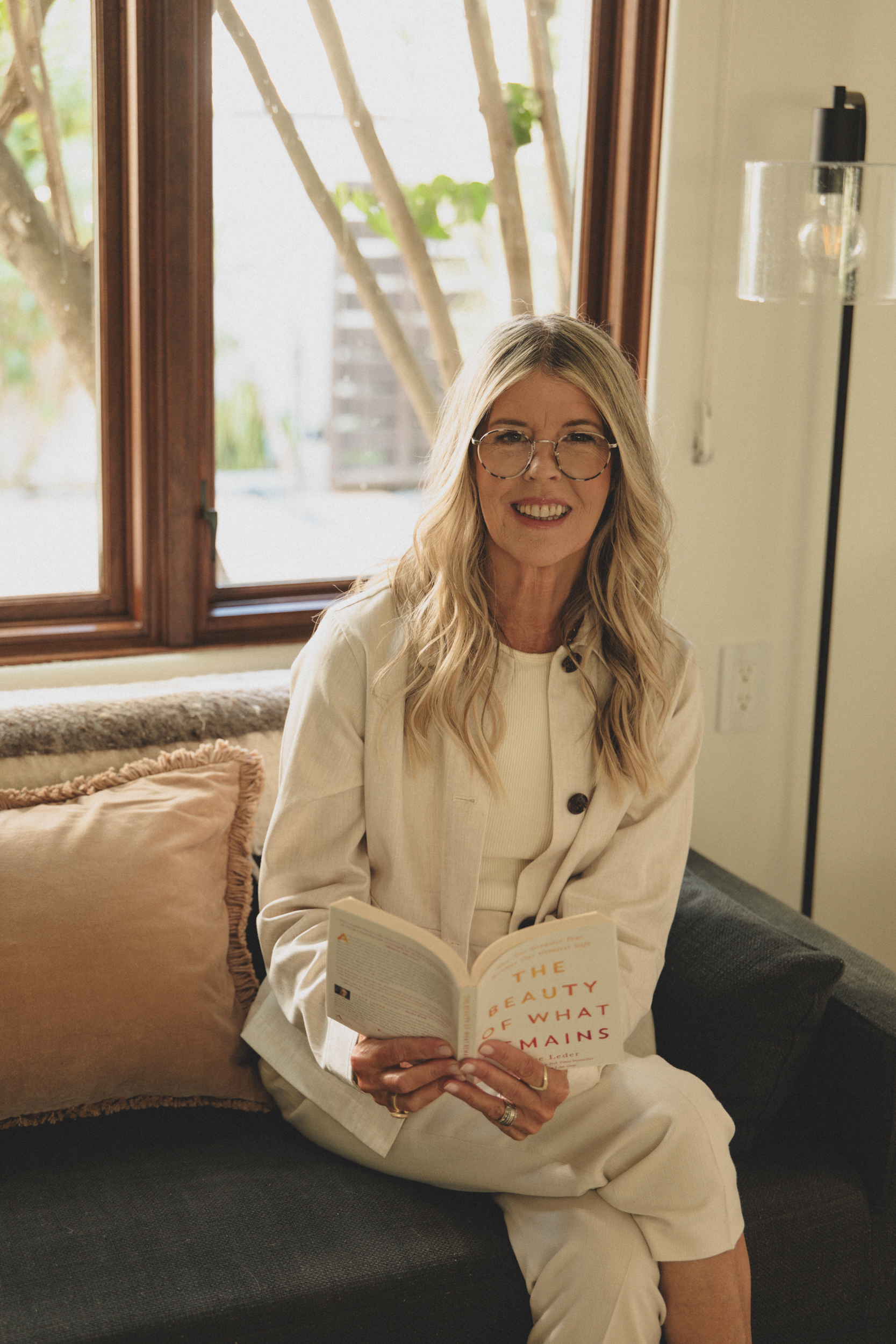 A woman with blonde hair and glasses, smiling, sitting on a black sofa inside a house by a window, reading a book titled 'The Beauty of What Remains'.