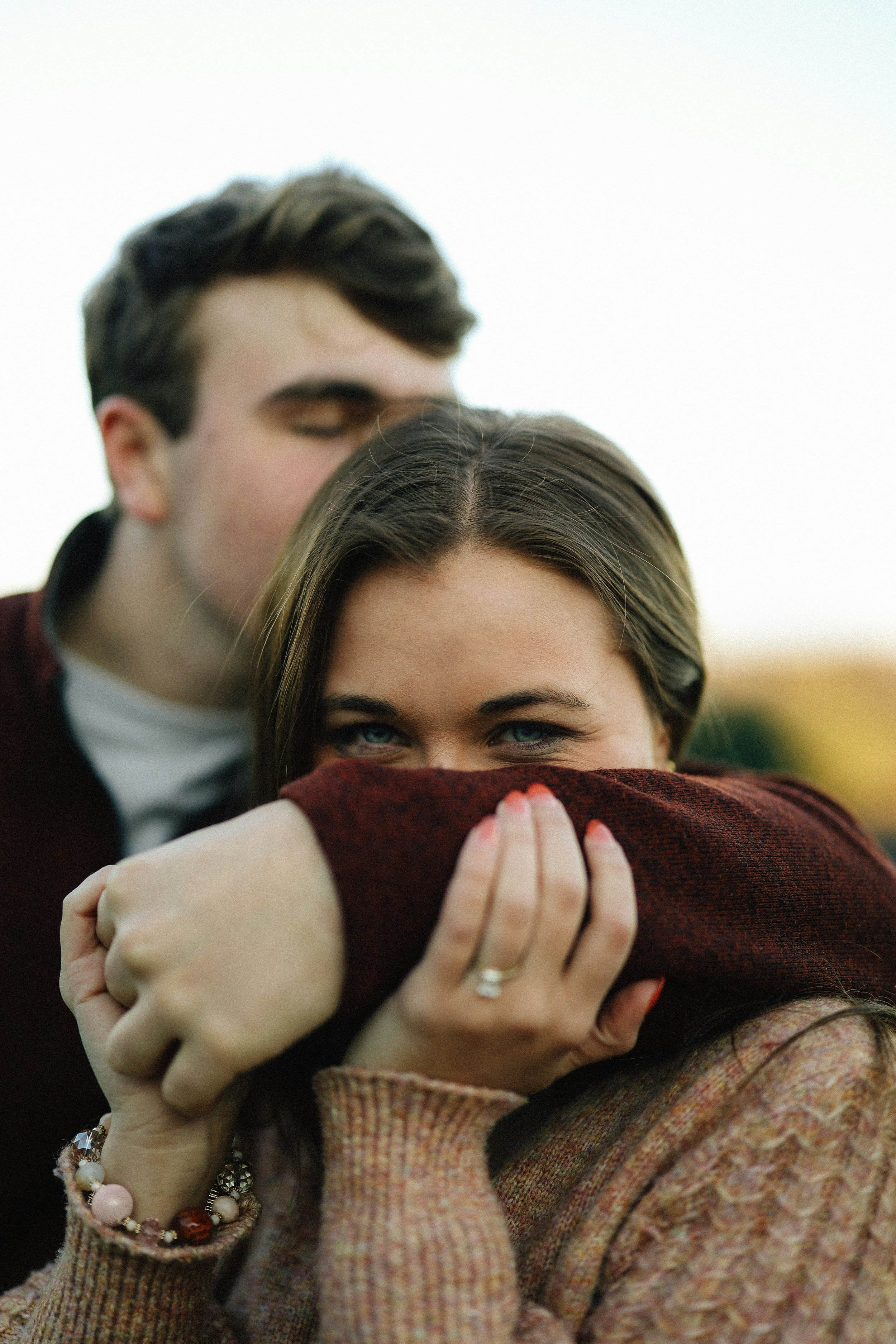 A close-up of two people embracing, with the woman's face partially visible and the man's face slightly blurred in the background. The woman has blue eyes and brown hair, and she is wearing a plaid jacket and jewelry on her wrist.