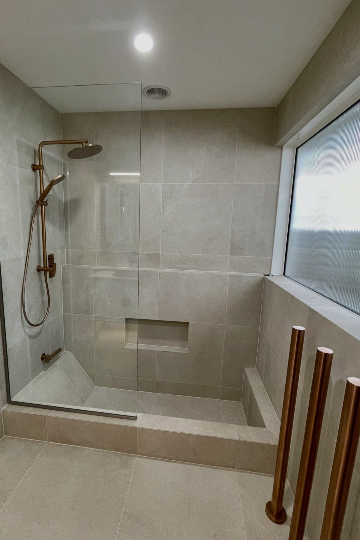 Modern shower with beige tiles, a built-in bench, a large frosted window, and a gold showerhead with a handheld shower.