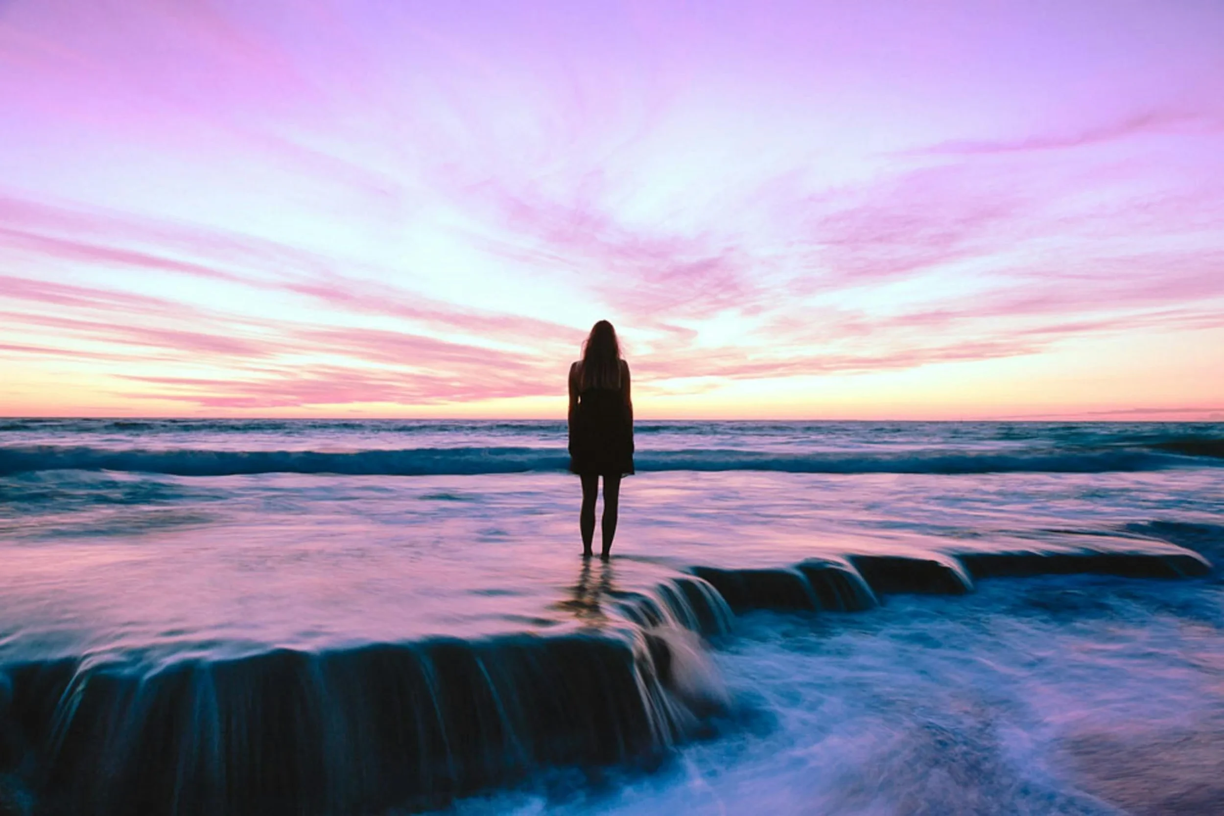 Woman standing peacefully in the ocean at sunset, symbolizing inner peace, alignment and embodiment - Desired Divinity