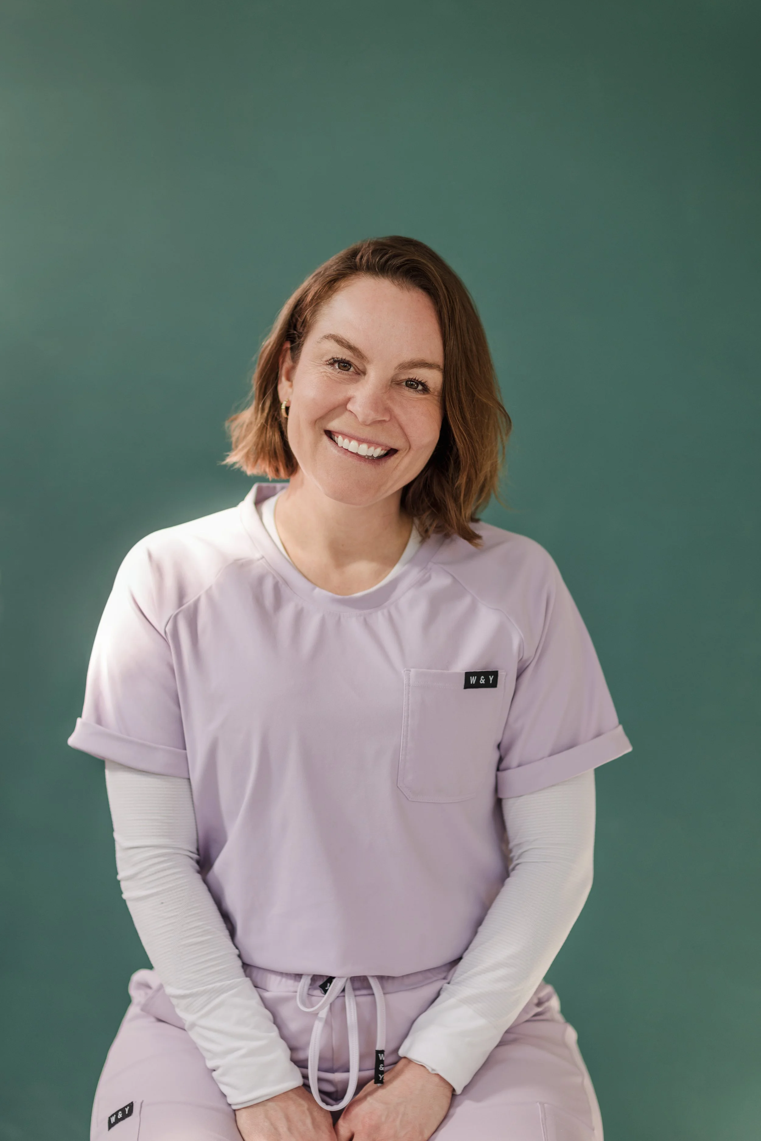 Une femme souriante avec des cheveux bruns courts, portant une tenue de travail violette avec des manches longues blanches en dessous, posant devant un fond vert