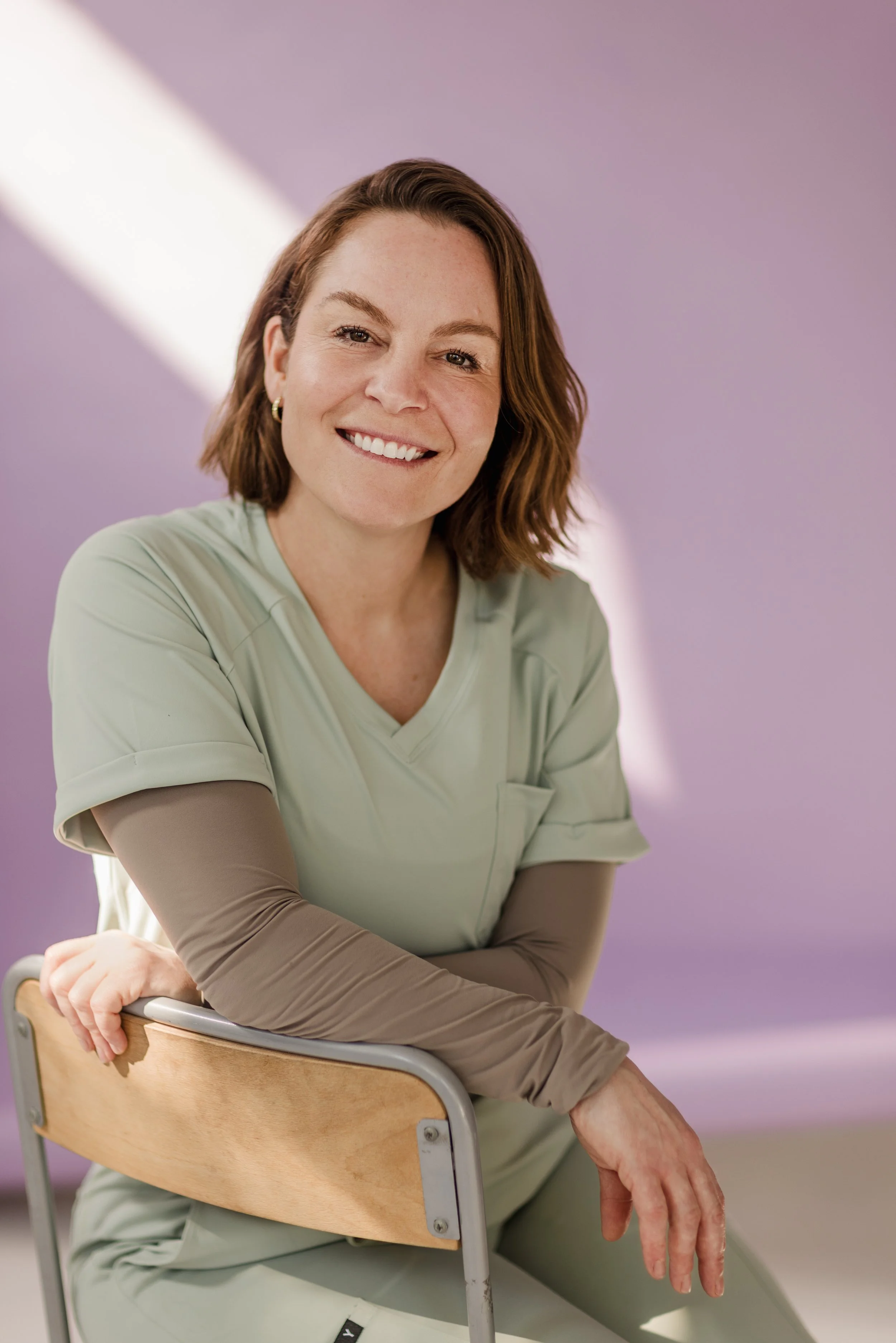 Une femme souriante avec des cheveux bruns courts, portant un pull couleur pastel, assise sur une chaise en bois. Elle a une main sur la chaise et l'autre suspendue, dans une pièce au mur violet clair.