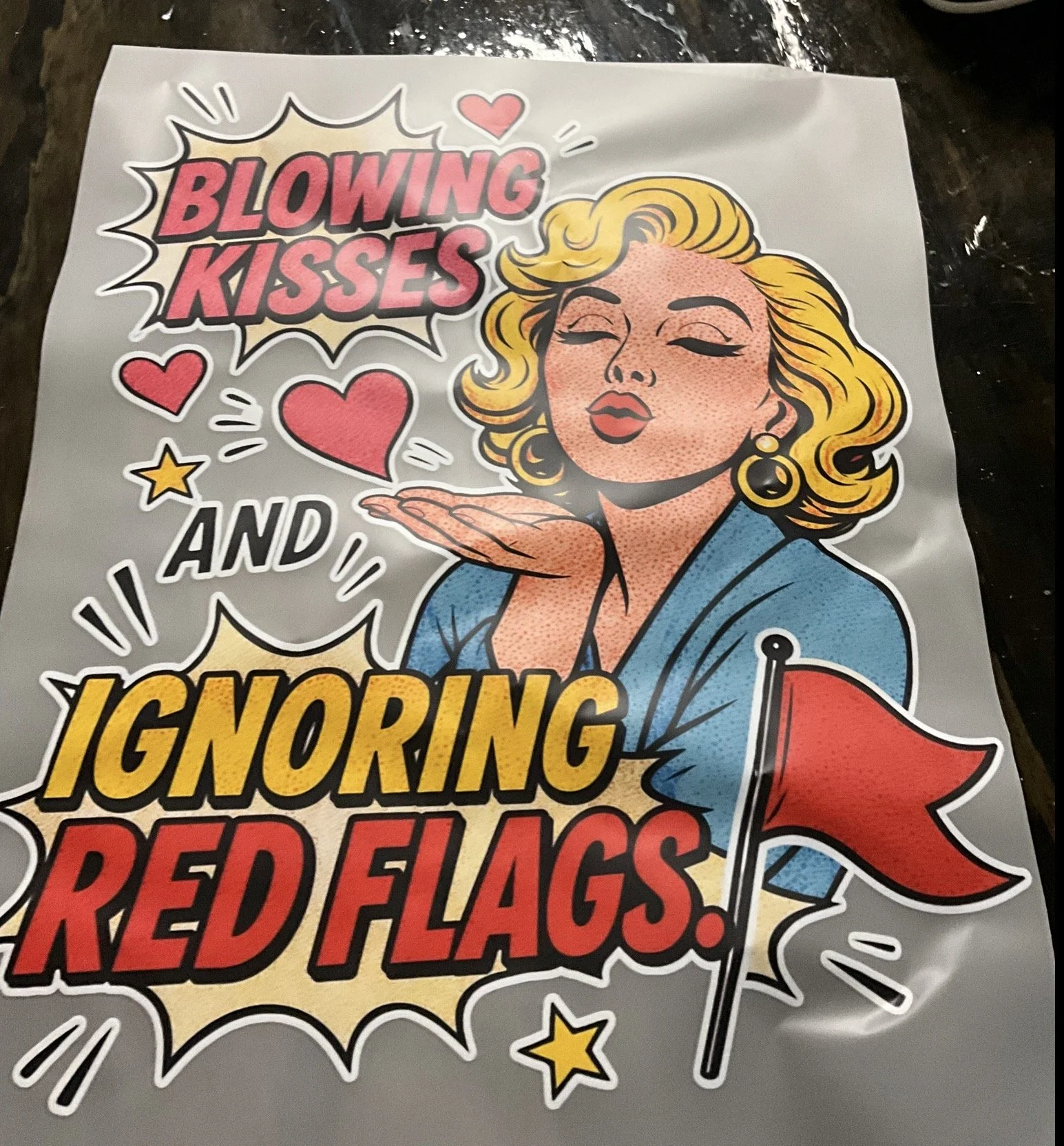 Pop art style illustration of a woman with blonde hair blowing a kiss, with hearts and stars around her, and the words 'Blowing kisses and ignoring red flags' in a comic book style.