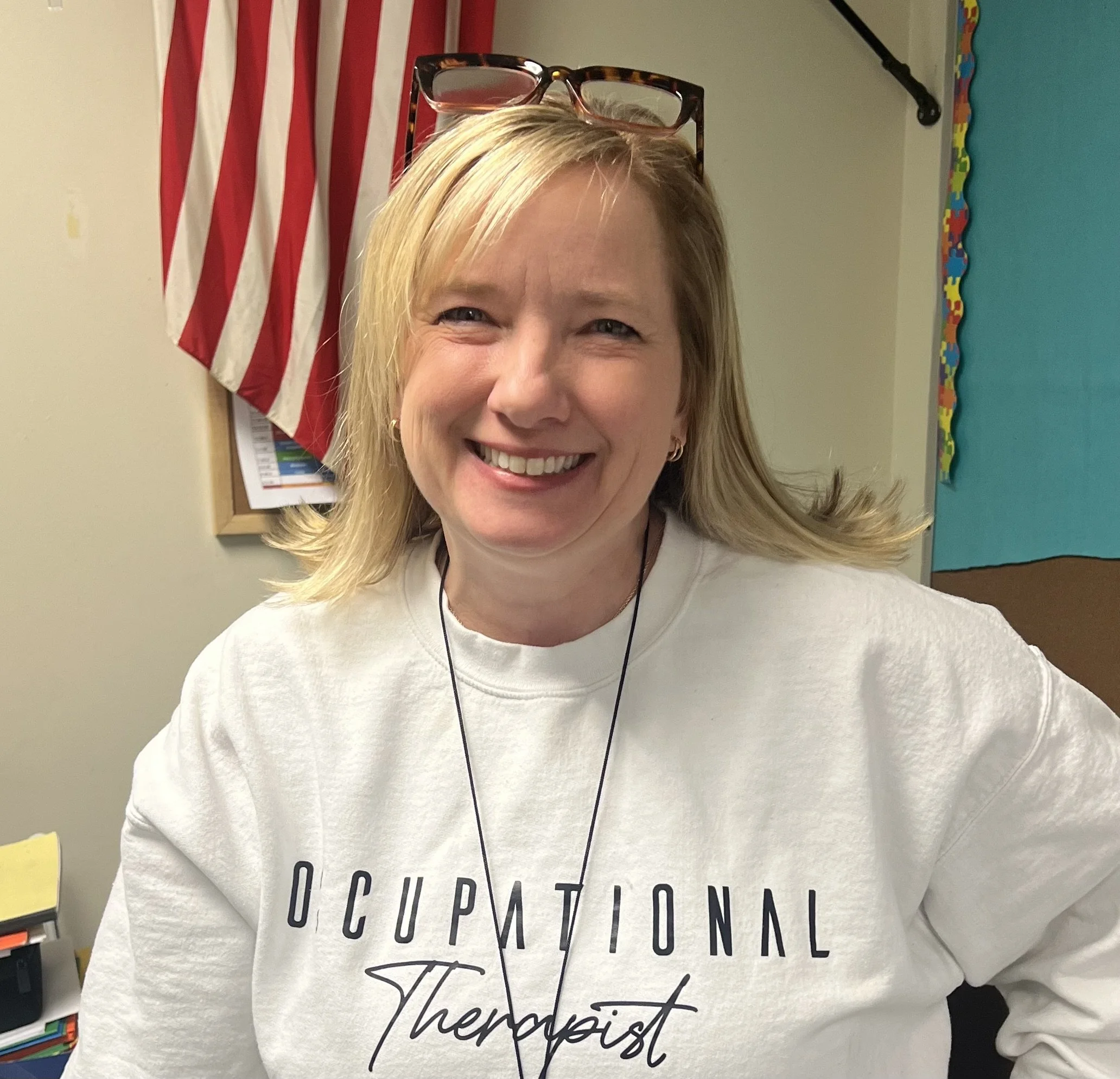 Close-up of a smiling woman with blonde hair, wearing glasses on her head and a white shirt with black text, standing in a classroom with an American flag and a colorful bulletin board in the background.