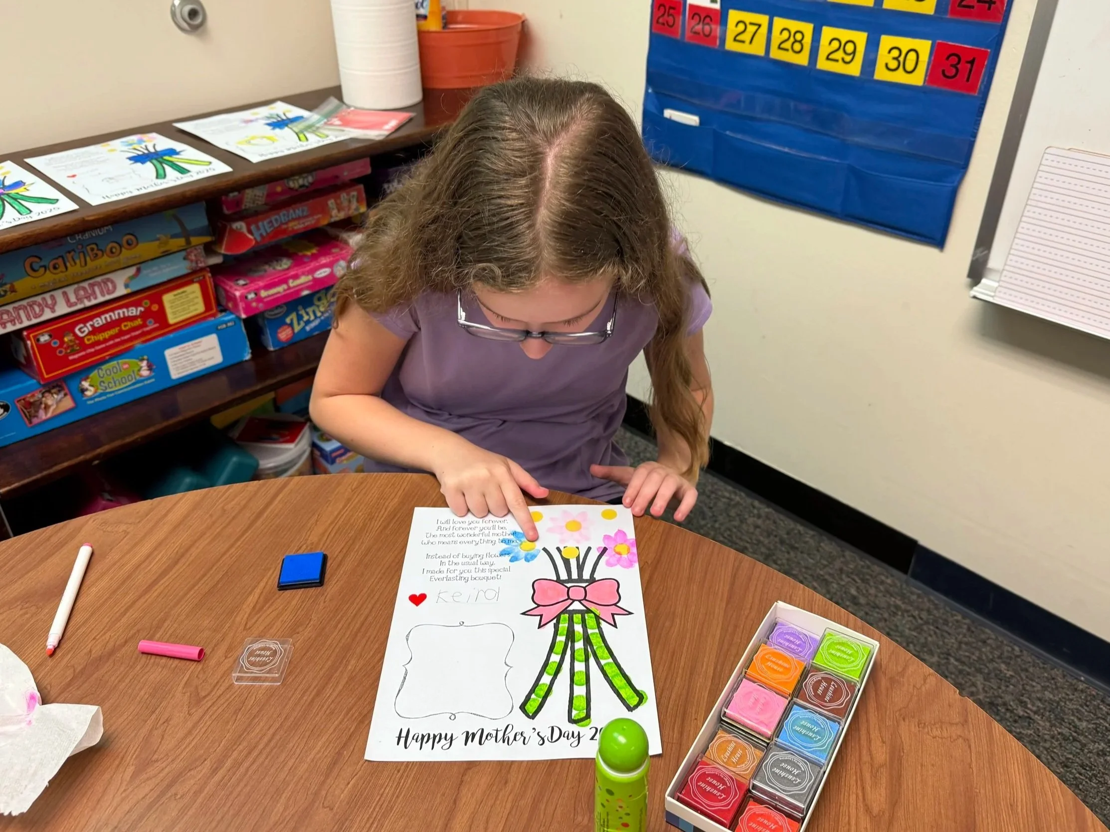 A young girl with glasses sitting at a table, coloring a Mother's Day card featuring a large flower with a pink bow, a red heart, and the words 'Happy Mother's Day' at the bottom. Various colored markers and ink pads are on the table.