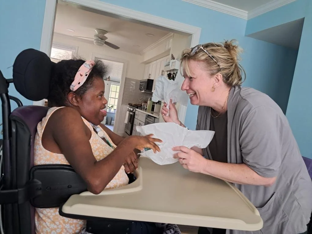 Two women smiling at each other, one of whom is in a wheelchair, inside a brightly lit home. The woman in the wheelchair has dark skin and curly hair, wearing a headband and sleeveless top. The other woman has light skin and blonde hair pulled back, wearing glasses and a light gray shirt, holding tissue paper.