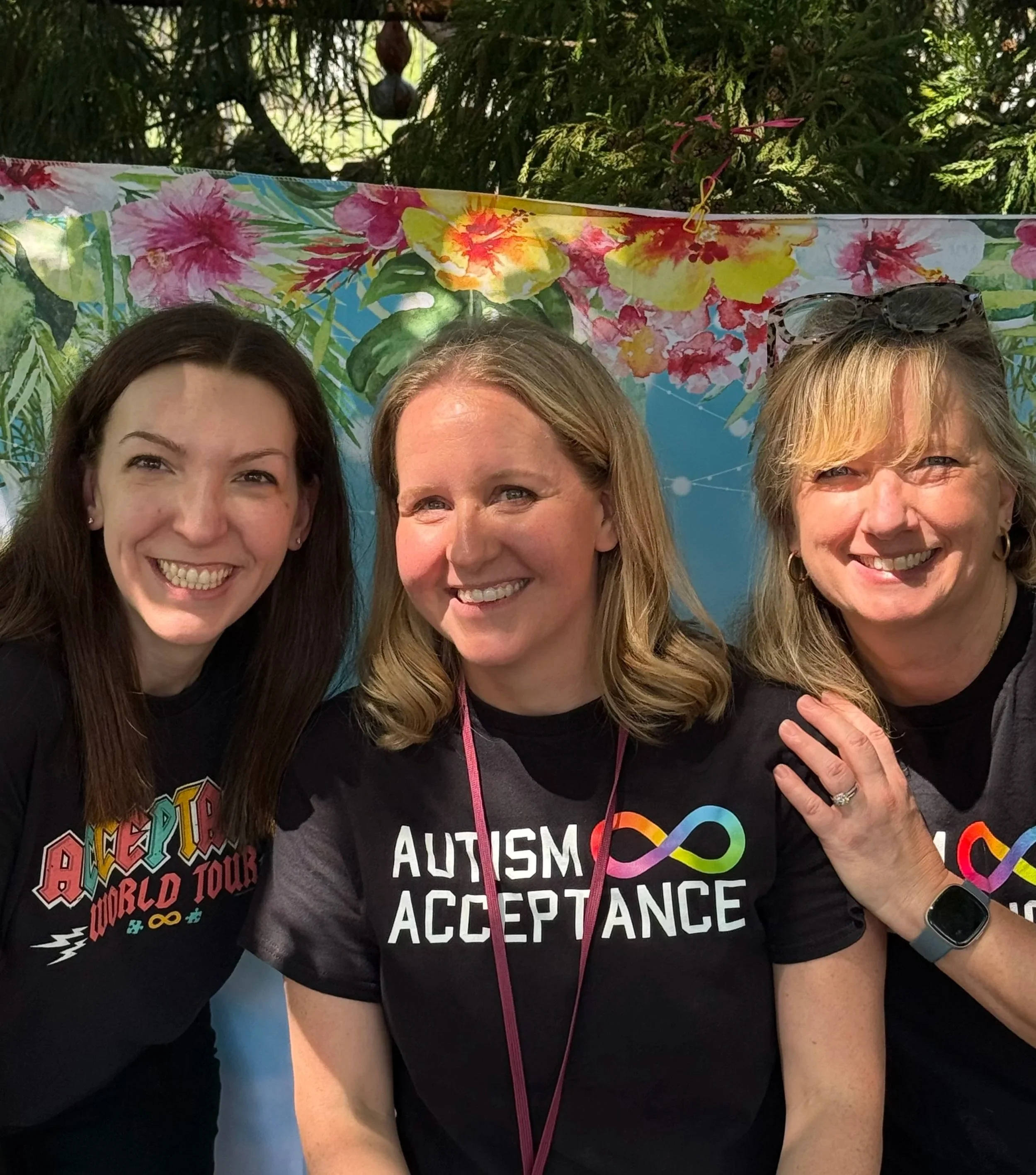 Three women smiling for a photo at an outdoor event with a colorful floral curtain in the background. One woman wears sunglasses on her head, another has curly hair, and the third has straight blonde hair. The middle woman is wearing a black shirt with the words 'Autism Acceptance' and a rainbow infinity symbol.