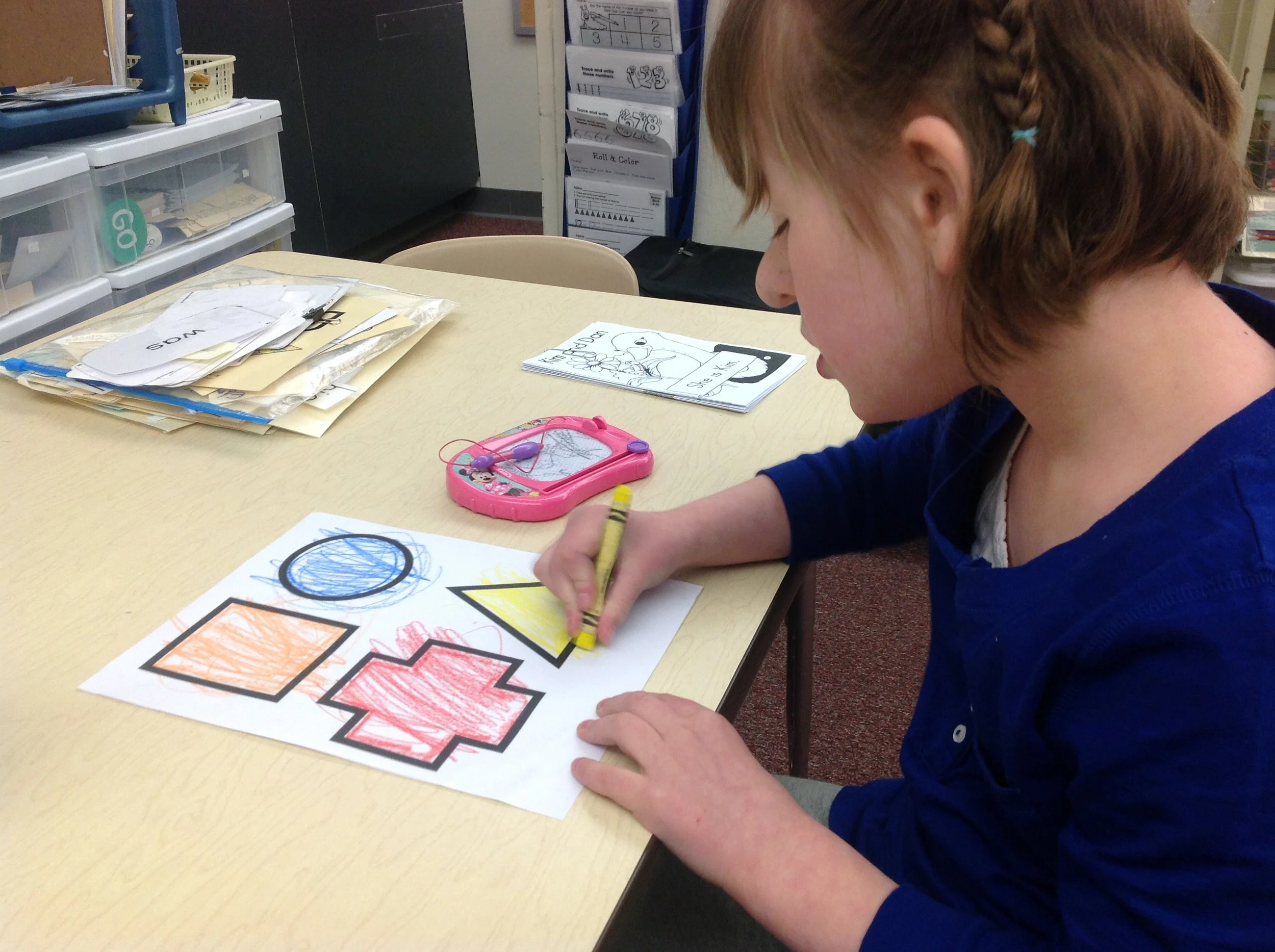 A young girl with braided hair sitting at a table doing coloring activities. She is using a yellow crayon to color a worksheet with different shapes, including circles and squares, and some coloring pages featuring characters like Mickey Mouse. The background shows educational supplies and organized folders in a classroom setting.