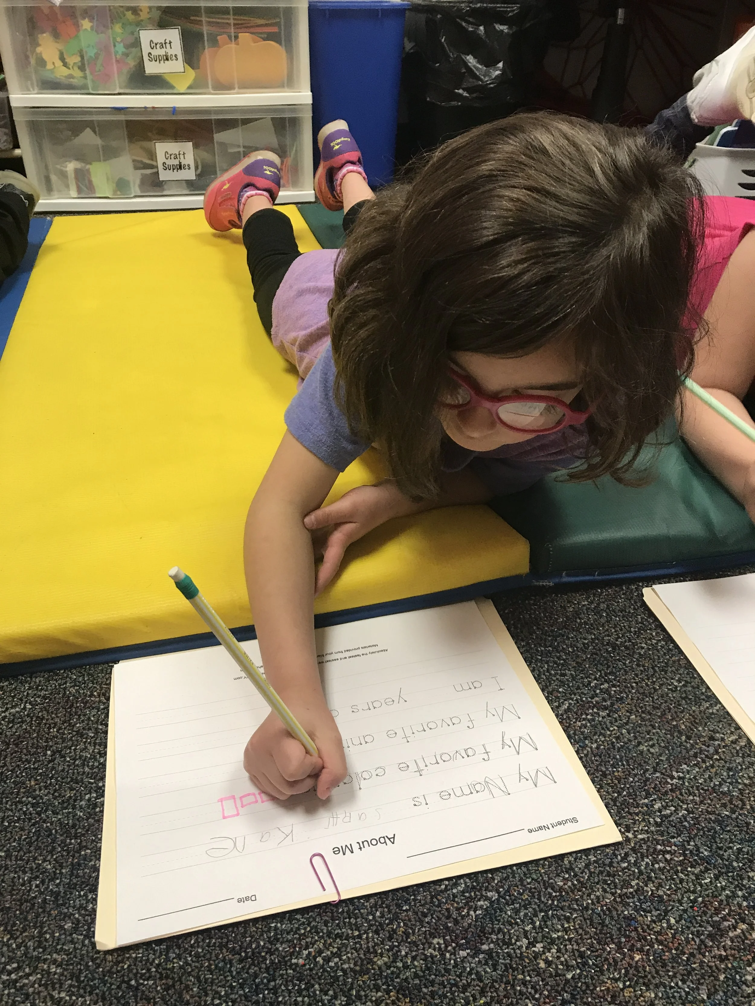 A young girl with glasses and curly hair is lying on her stomach on a yellow mat, writing on a large sheet of lined paper with a green pencil. The paper has a list of sentences with checkboxes. In the background, there are plastic storage drawers labeled 'Craft Supplies' and other classroom items.
