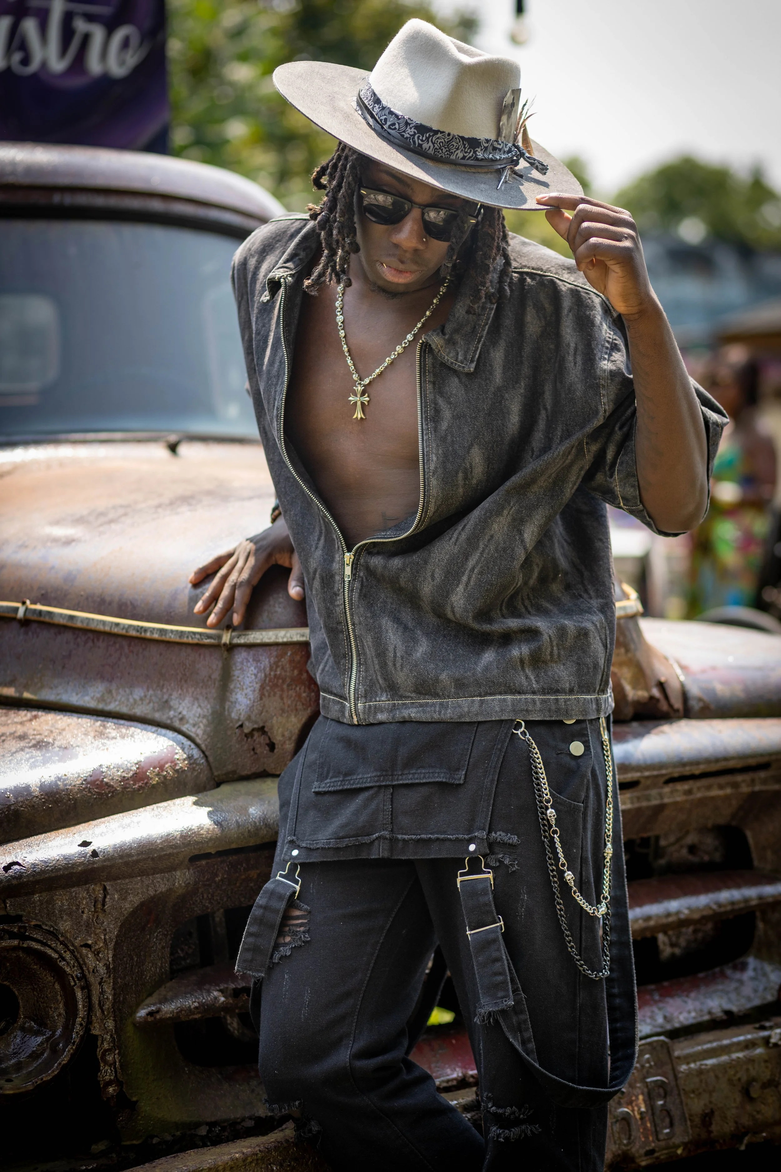 A person with dreadlocks wearing a wide-brimmed hat, sunglasses, a gold chain, a zip-up black denim jacket, and ripped black jeans with chains, standing next to an old, rusty truck outdoors.