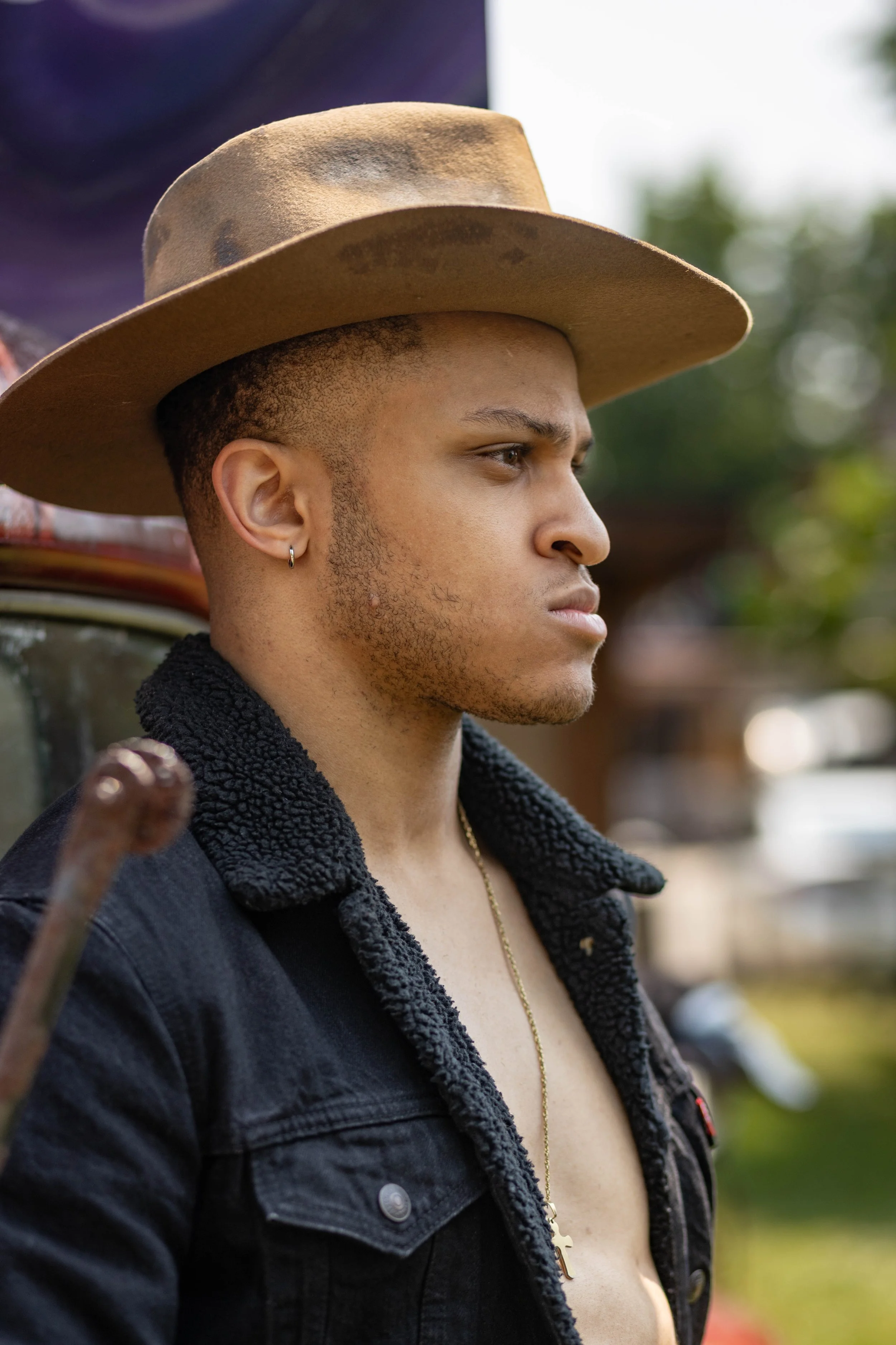 A young man with a clipper haircut, wearing a tan cowboy hat and a black jacket with a fleece collar, is looking to the side outdoors. He has a small hoop earring in his left ear and a gold cross necklace visible on his chest.