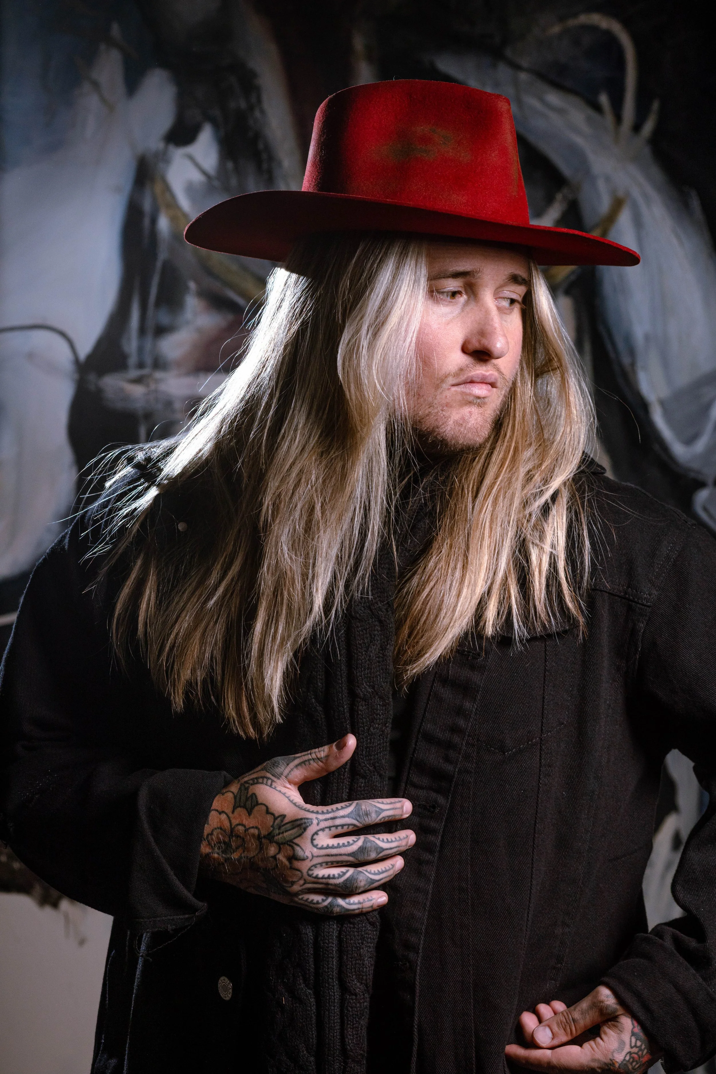A man with long blond hair and tattoos on his hand wearing a red wide-brimmed hat, black jacket, and black shirt, standing in front of a dark, artistic background.