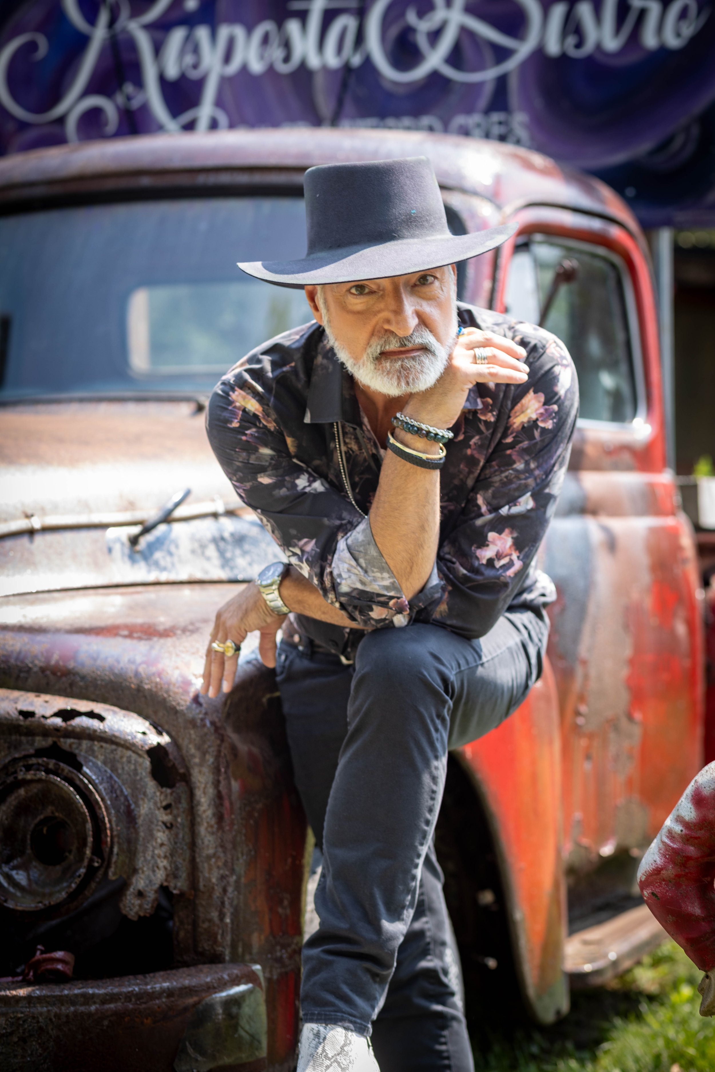 A man with a grey beard wearing a large black hat and floral shirt, leaning on a rusty, old red truck, with one hand on his chin and a contemplative expression.