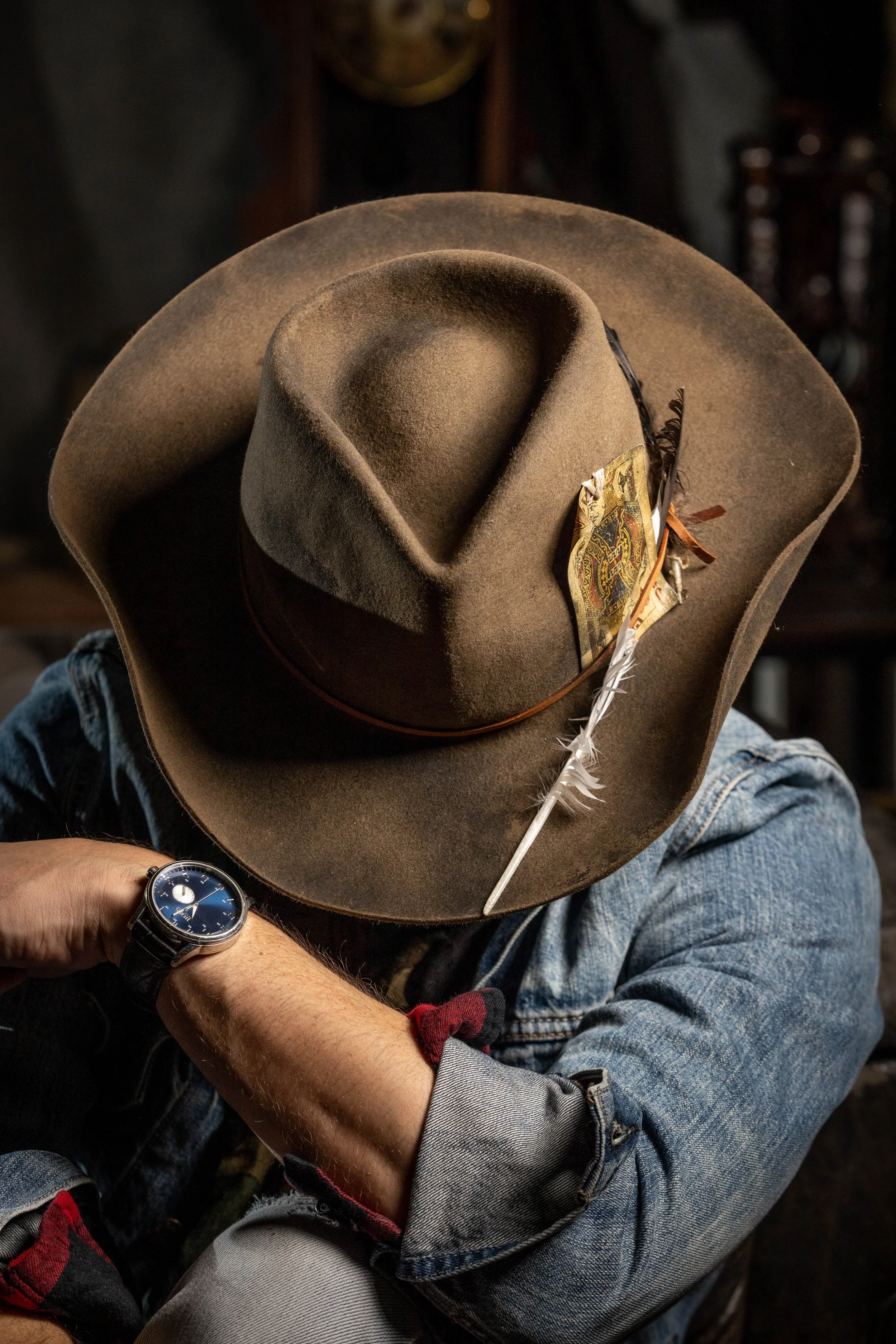 A person wearing a denim jacket and jeans, with a watch on their wrist, holds a wide-brimmed brown felt cowboy hat decorated with a feather and a paper band.