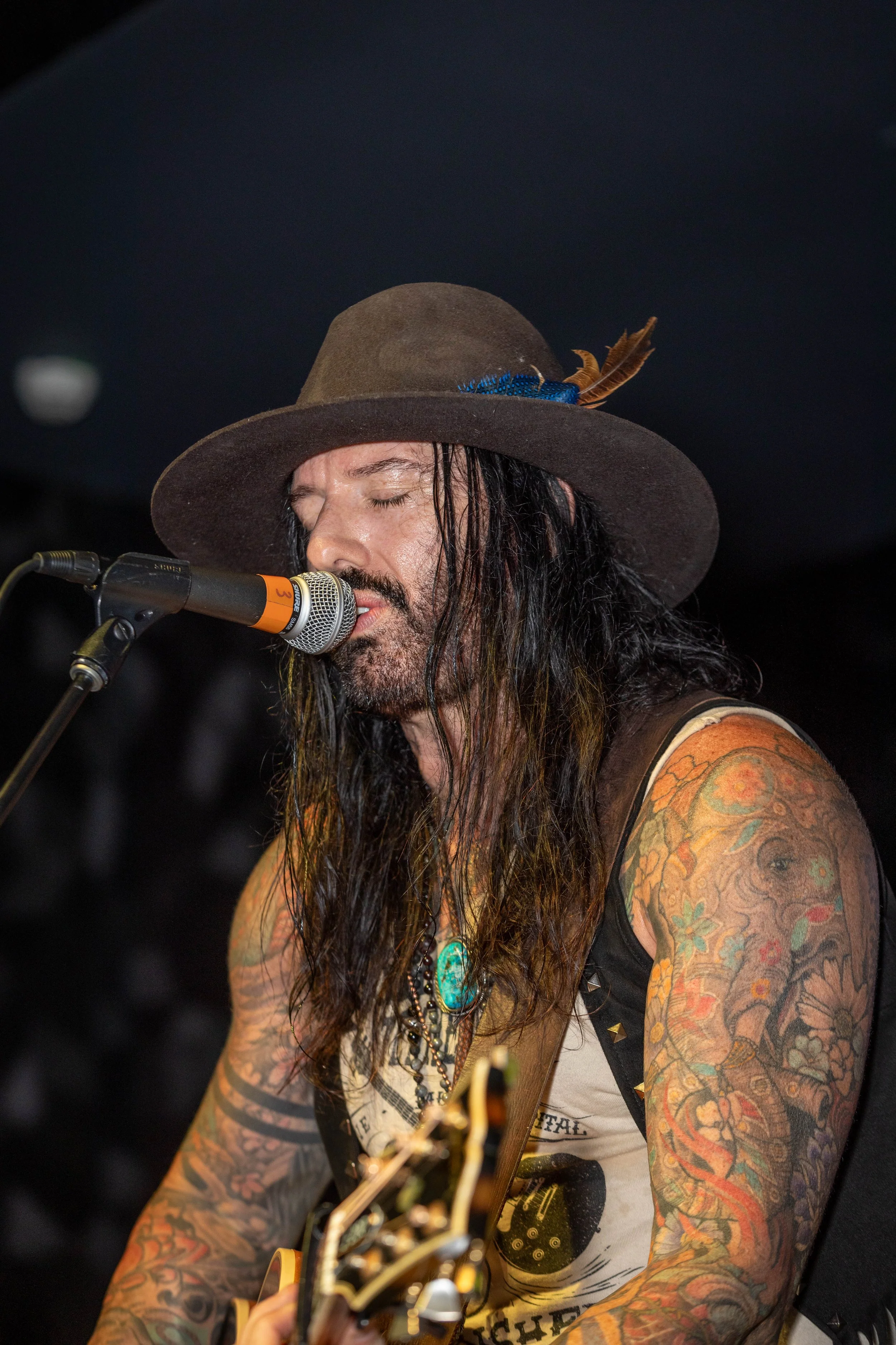 A man with long dark hair and tattoos on his arms is singing into a microphone while playing an acoustic guitar. He wears a gray hat with a feather and a sleeveless shirt with artwork. The background is dark and out of focus.