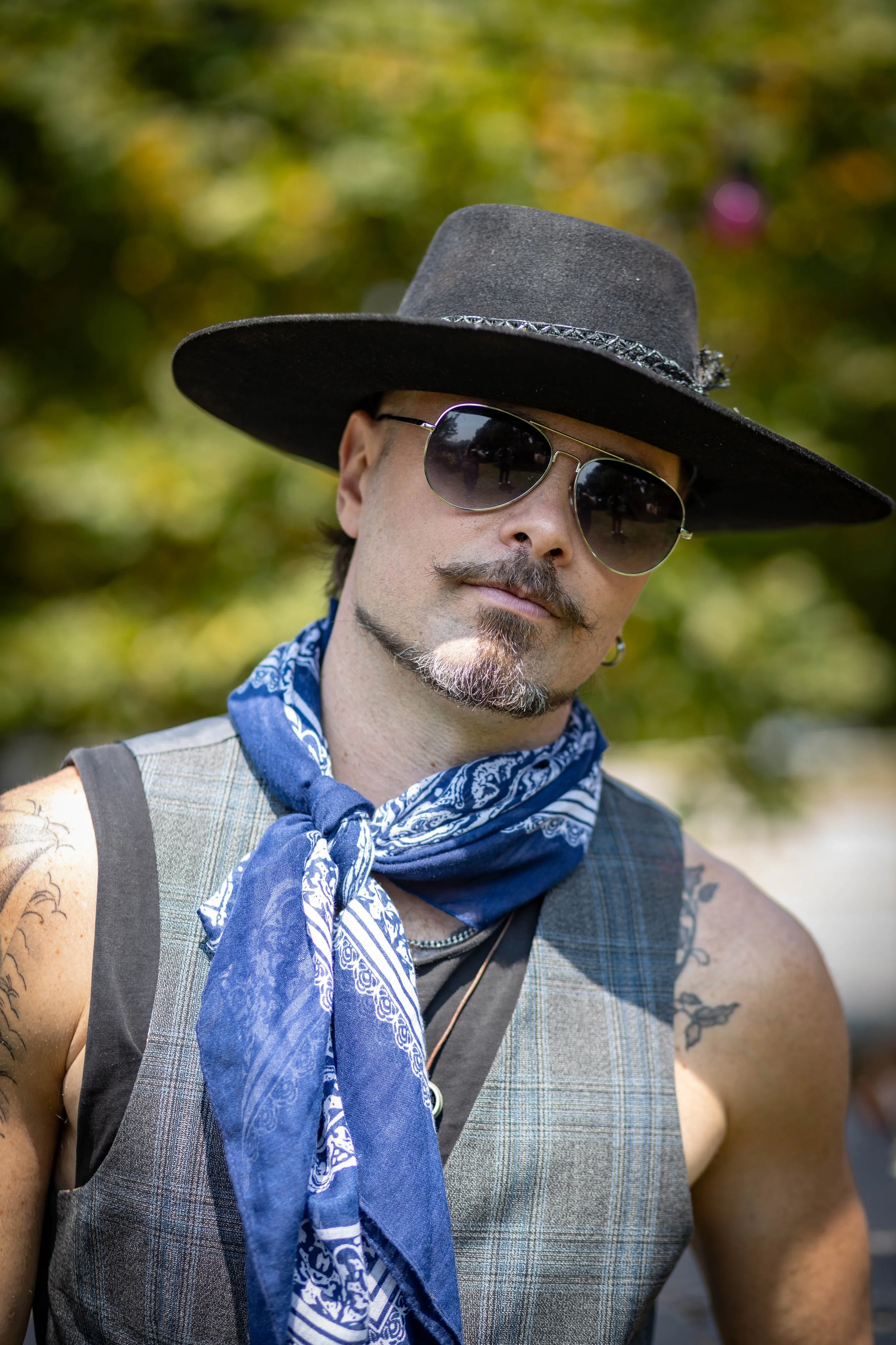 Man wearing a wide-brimmed black hat, aviator sunglasses, a blue bandana, and a sleeveless shirt outdoors with blurred trees in the background.