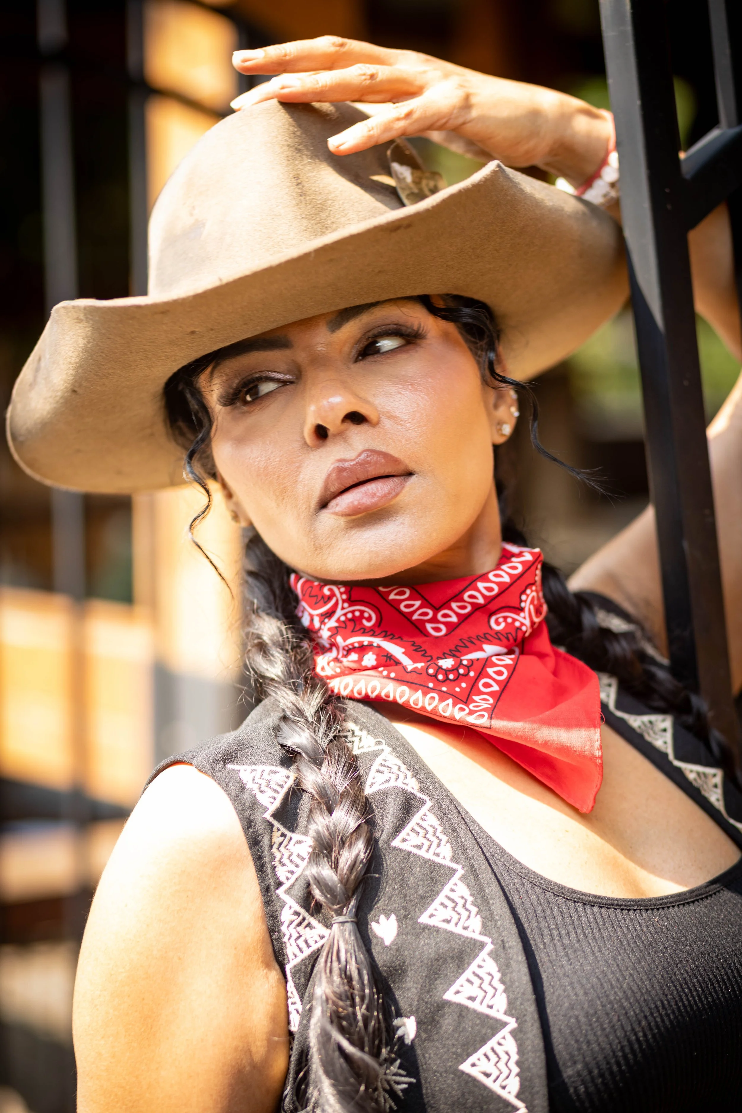 A woman with braided hair, wearing a large beige cowboy hat, a red bandana around her neck, and a black tank top with white geometric patterns.