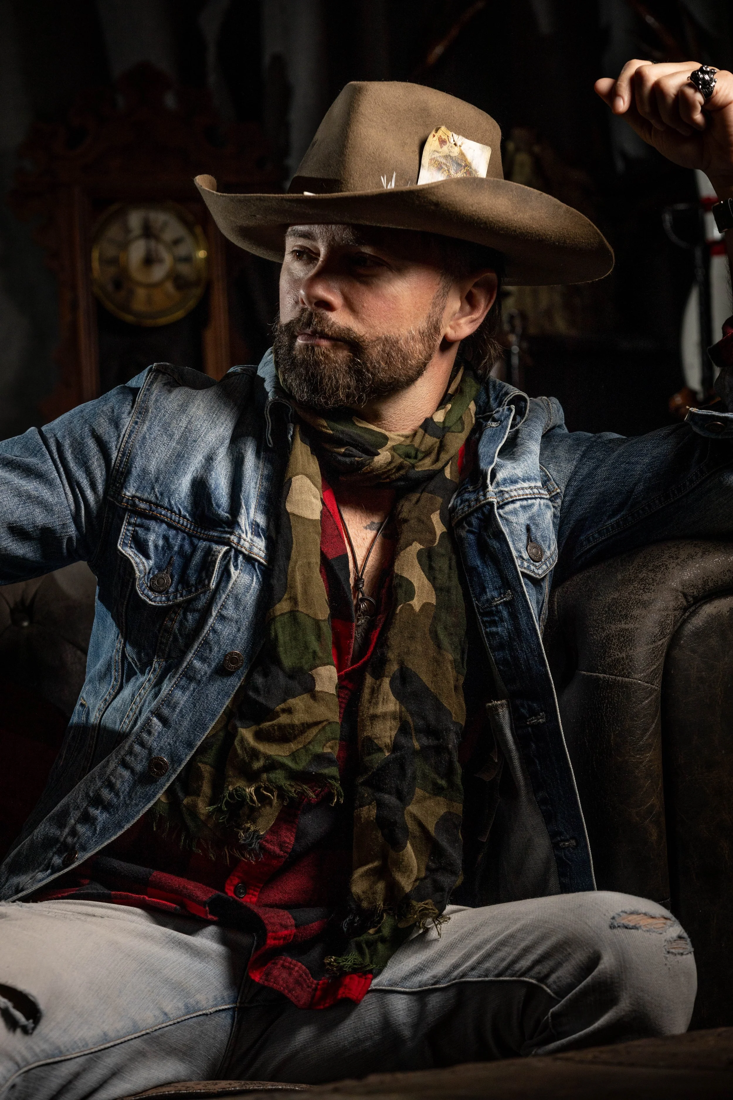 A man with a beard wearing a wide brown hat, a camouflage scarf, a denim jacket, and ripped jeans, sitting and flexing his arm.
