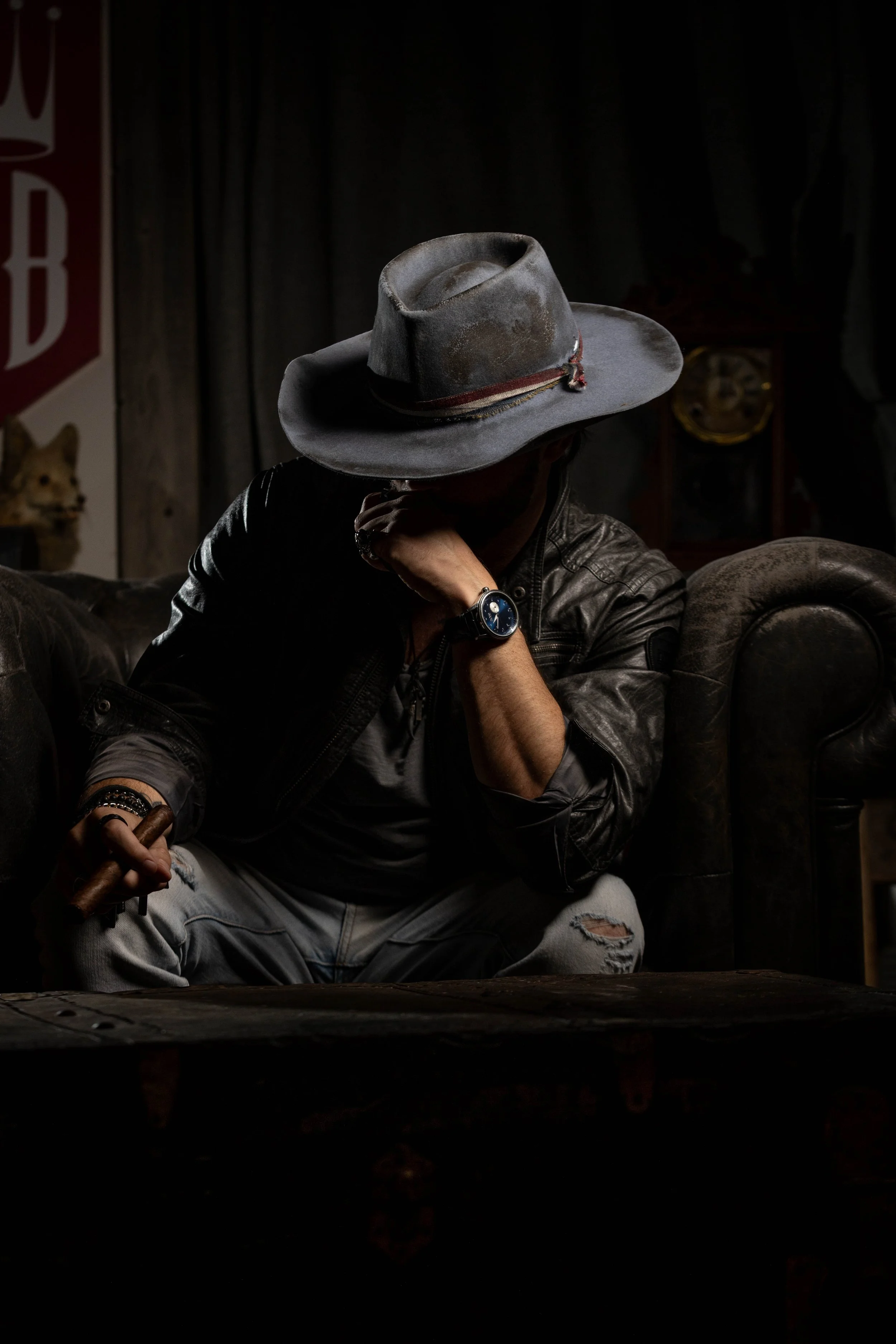 A person wearing a wide-brimmed hat sitting in a dark room, resting their chin on their hand with a cigar in their other hand, dressed in a leather jacket and ripped jeans, with a vintage clock and dog poster in the background.