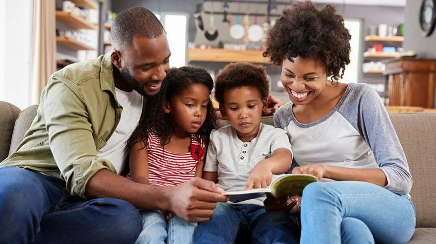 Family of four, three children and one adult female, sitting on a couch and reading a book together in a cozy living room.