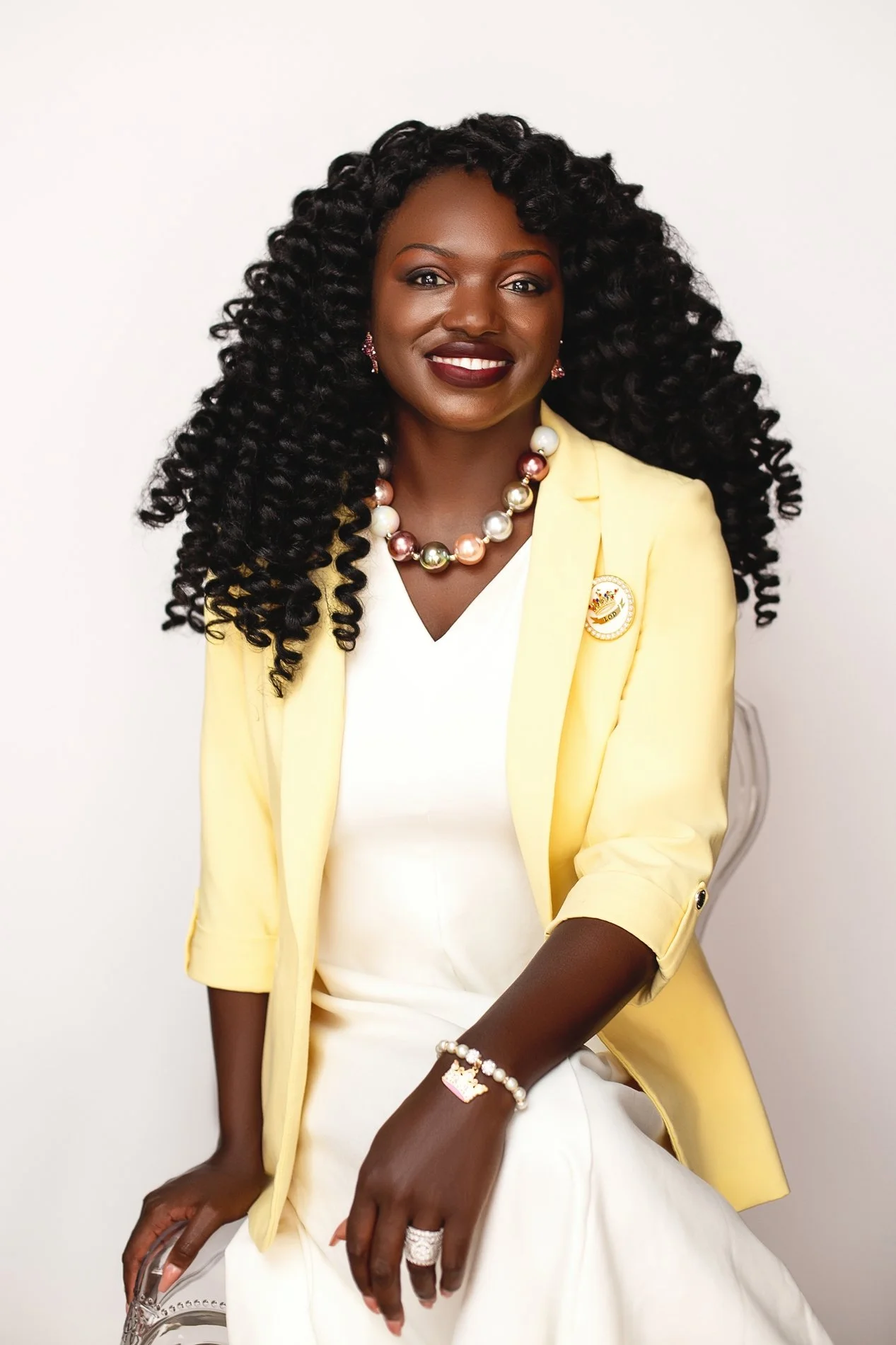 A woman with dark skin, long curly hair, wearing a white dress, yellow blazer, and colorful jewelry, sitting on a transparent chair against a plain white background.