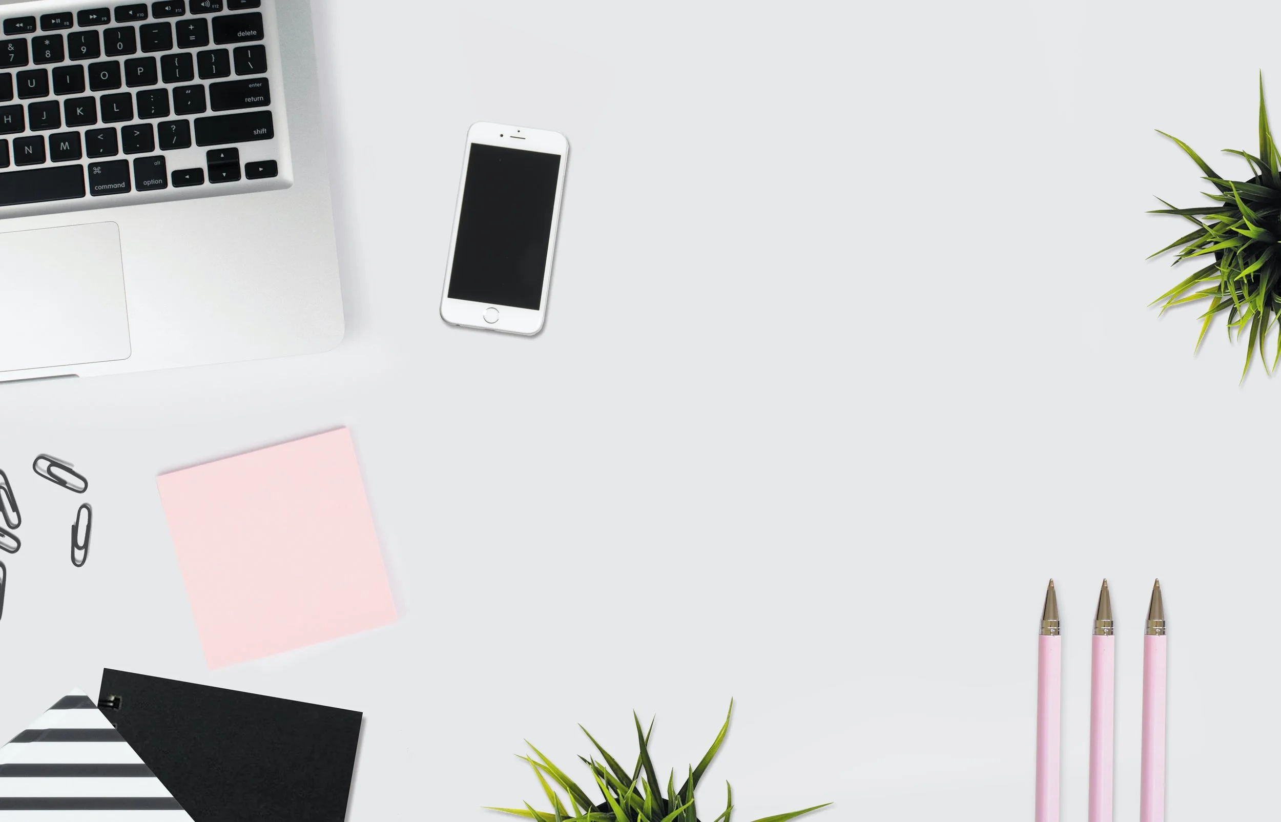Flat lay of a workspace with a silver laptop, an iPhone, a small potted plant, pink pens, paper clips, a black notebook, a pink sticky note, and striped paper on a white surface.
