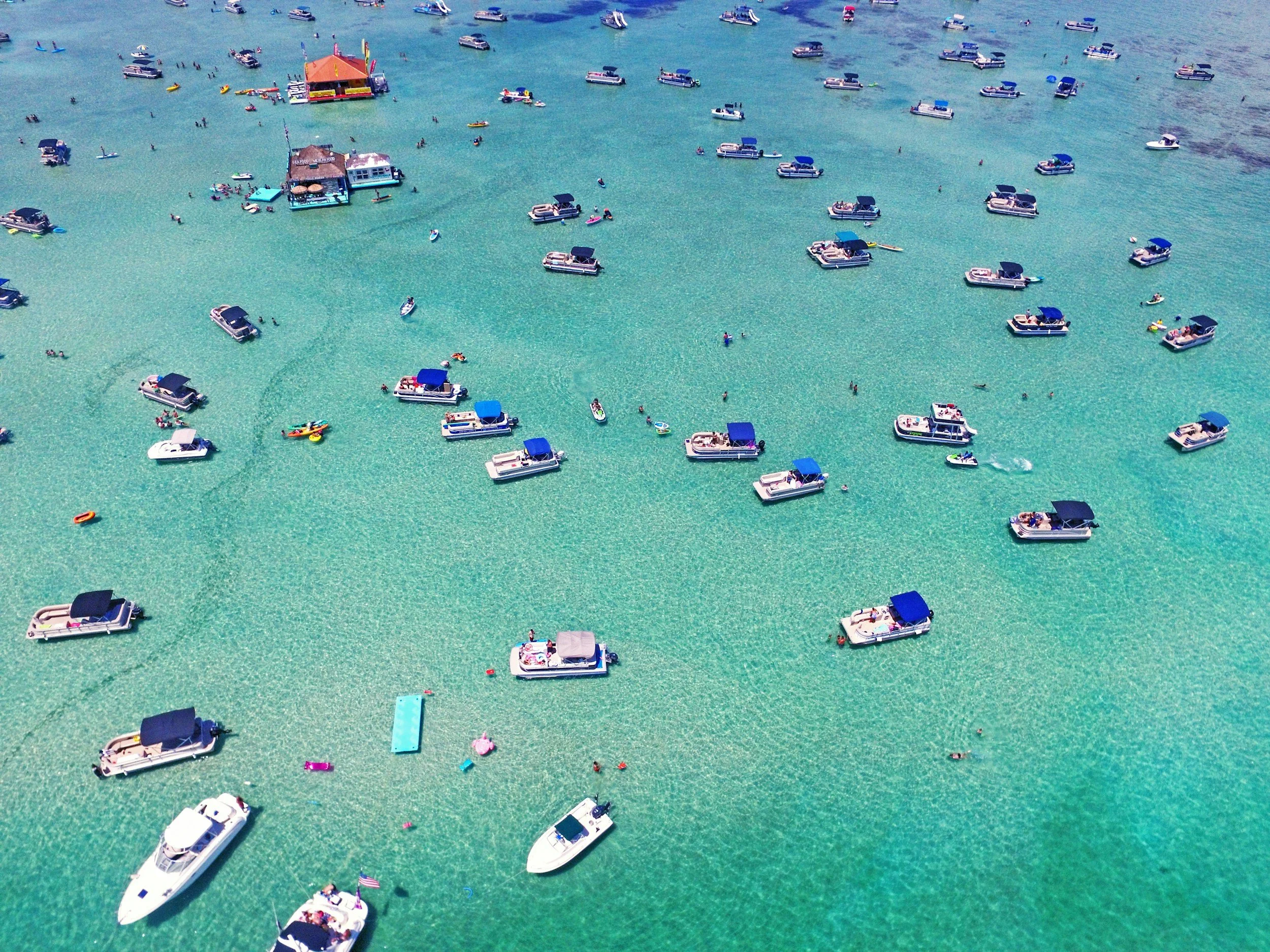 An aerial view of a crowded marina with various boats, including speedboats and pontoon boats, floating on clear turquoise water. Some boats have blue canopies and there are people swimming and enjoying the water.