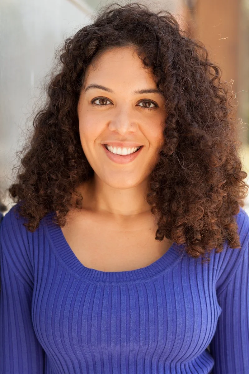 A woman with curly brown hair smiling, wearing a blue ribbed long-sleeve top, outdoors with blurred background.