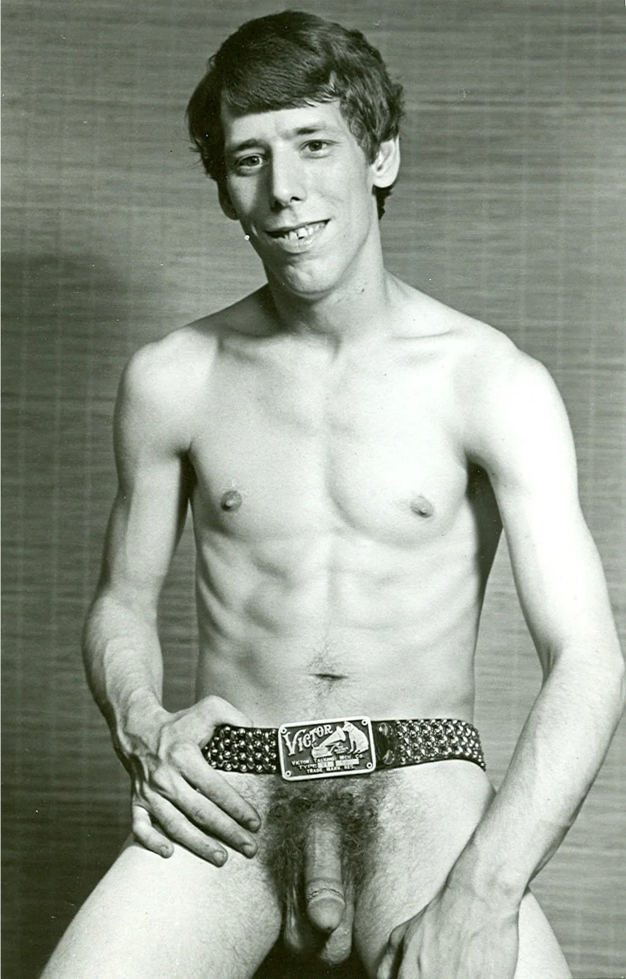 Black and white photo of lamp maker and hustler John Klar with a slim build, short hair, smiling, wearing a studded belt, and posing without clothing, against a plain background.