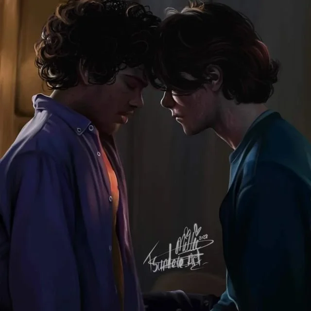 Two young men with curly hair and glasses lean their foreheads together in an emotional moment indoors, dimly lit.