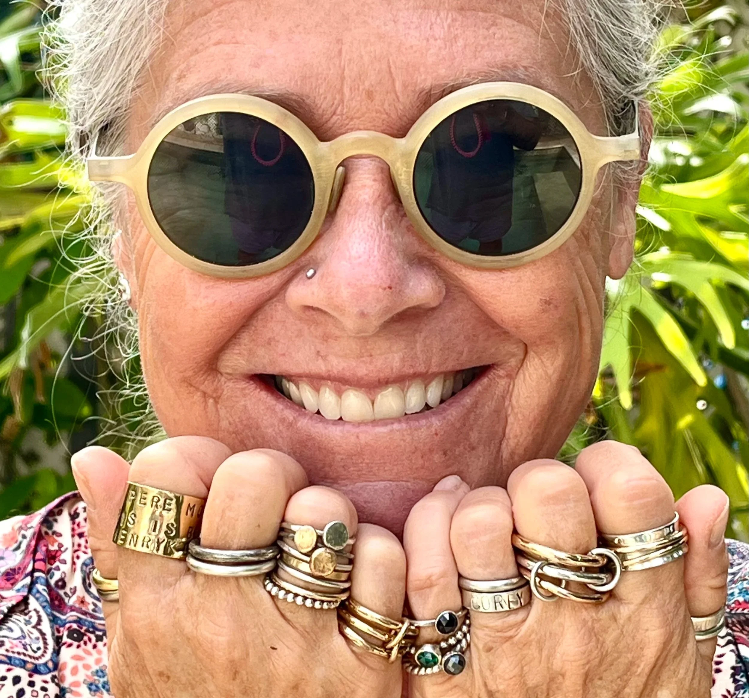 Close-up of a smiling woman with a band of white hair visible, surrounded by green foliage. She has several rings on her fingers and bracelets on her wrist, and she is holding her hands up near her face.