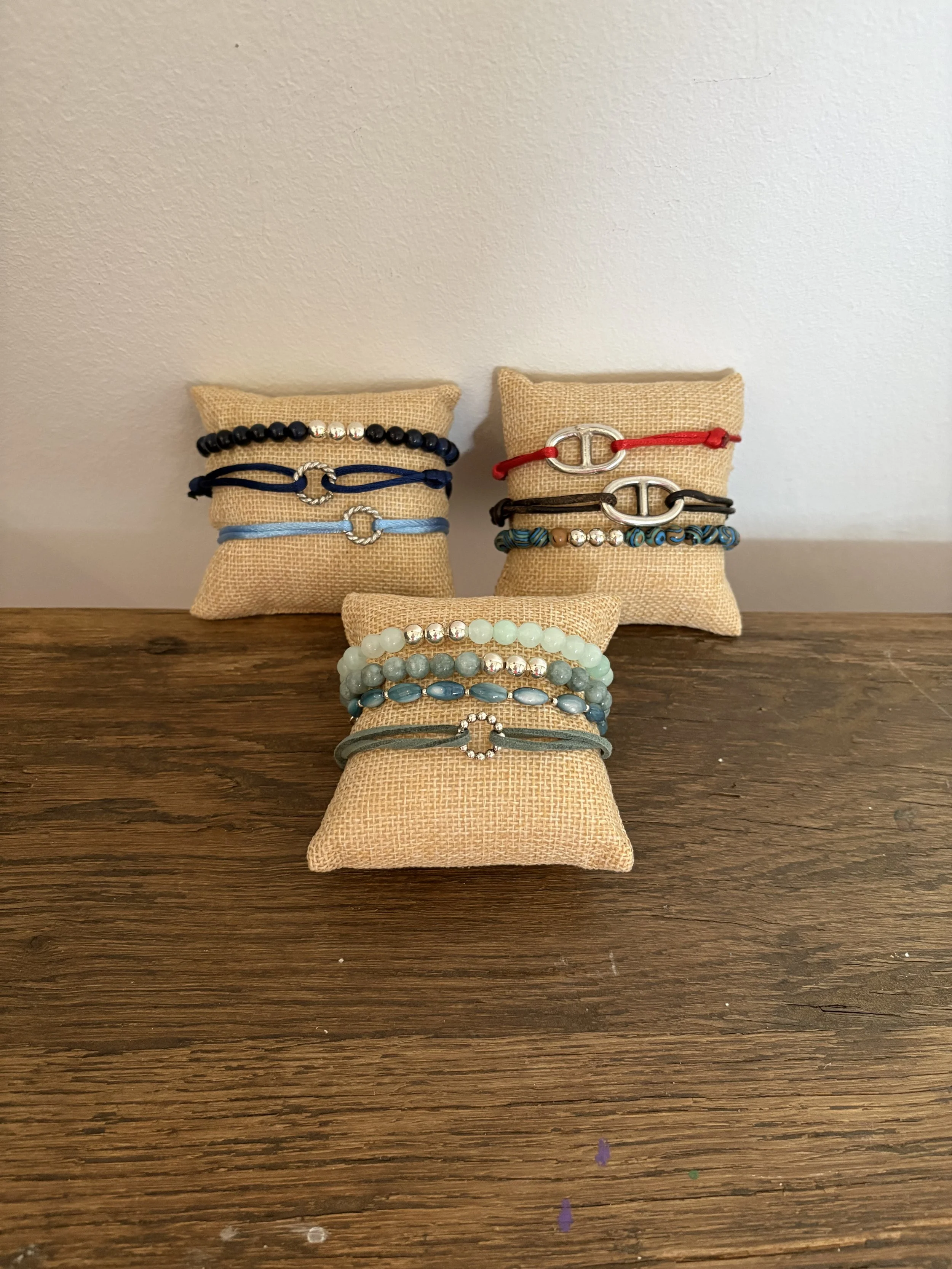 Display of eight bracelets on small burlap pillows, arranged on a wooden surface against a plain wall.