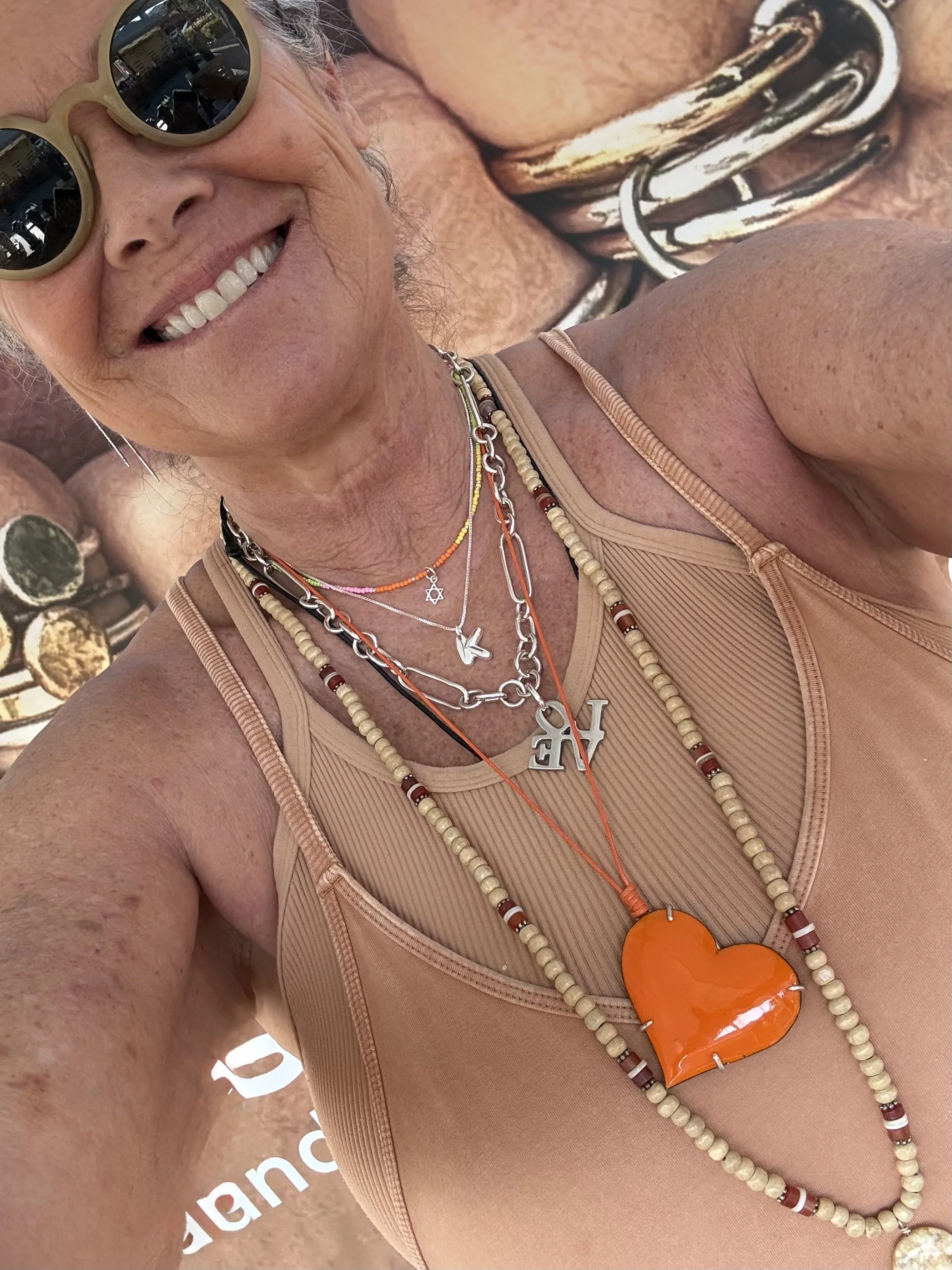 A woman smiling wearing sunglasses, a beige tank top, and multiple layered necklaces including a star of David and a large orange heart-shaped pendant.