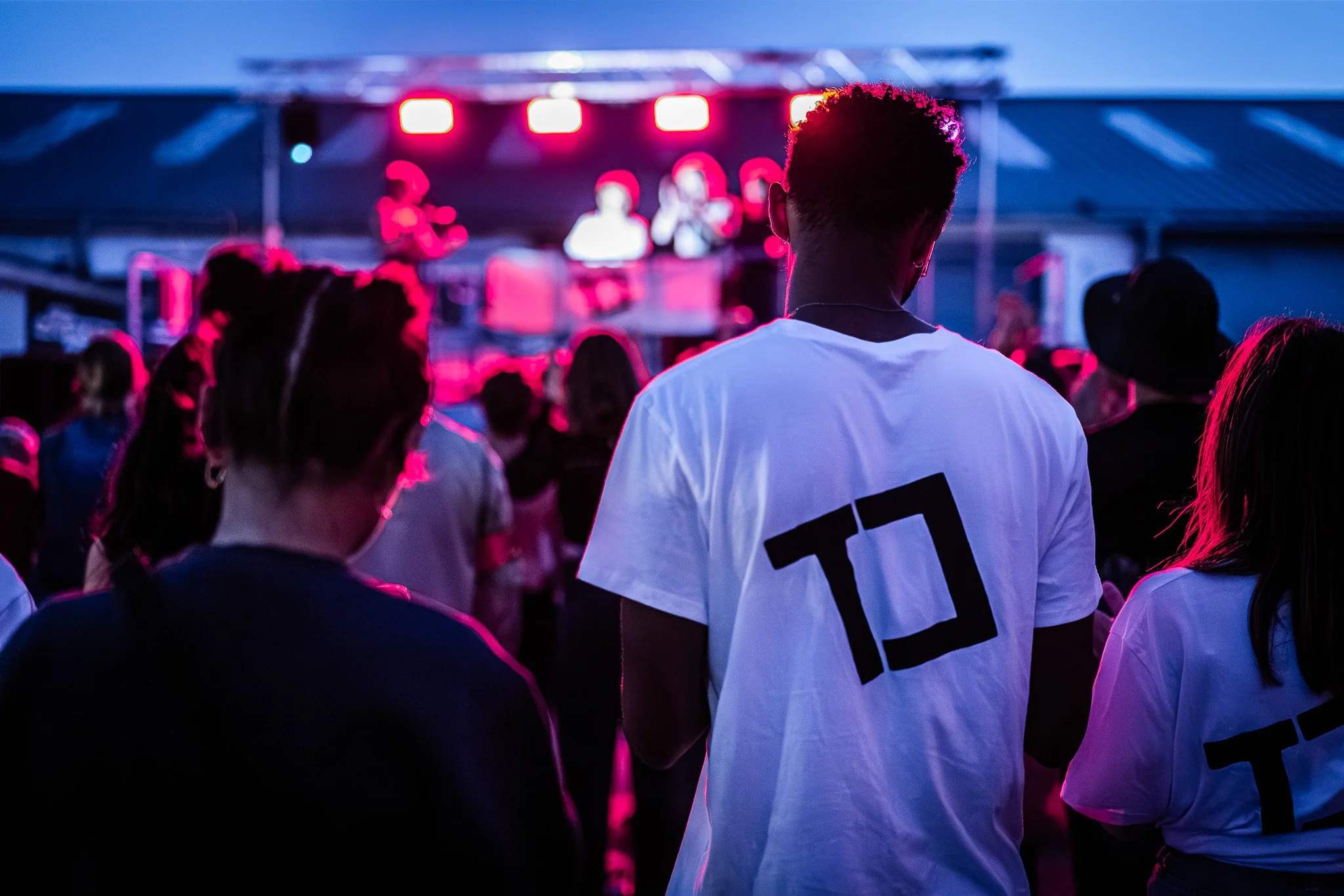 Jeune homme avec des cheveux courts en mouvement, portant un t-shirt blanc avec un numéro noir à l'arrière, regardant un spectacle ou un événement lors d'une soirée avec des lumières rouges en arrière-plan, entouré d'une foule.