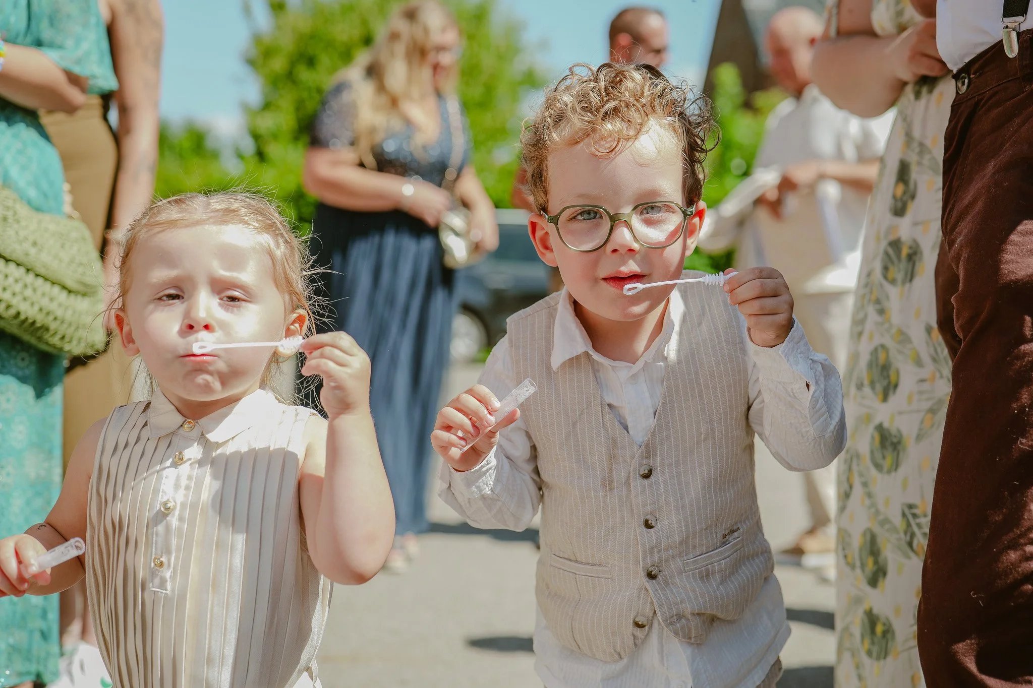 Deux jeunes enfants, une fille et un garçon, qui soufflent dans des bulles de savon lors d'une activité en plein air par une journée ensoleillée, entourés d'adultes.