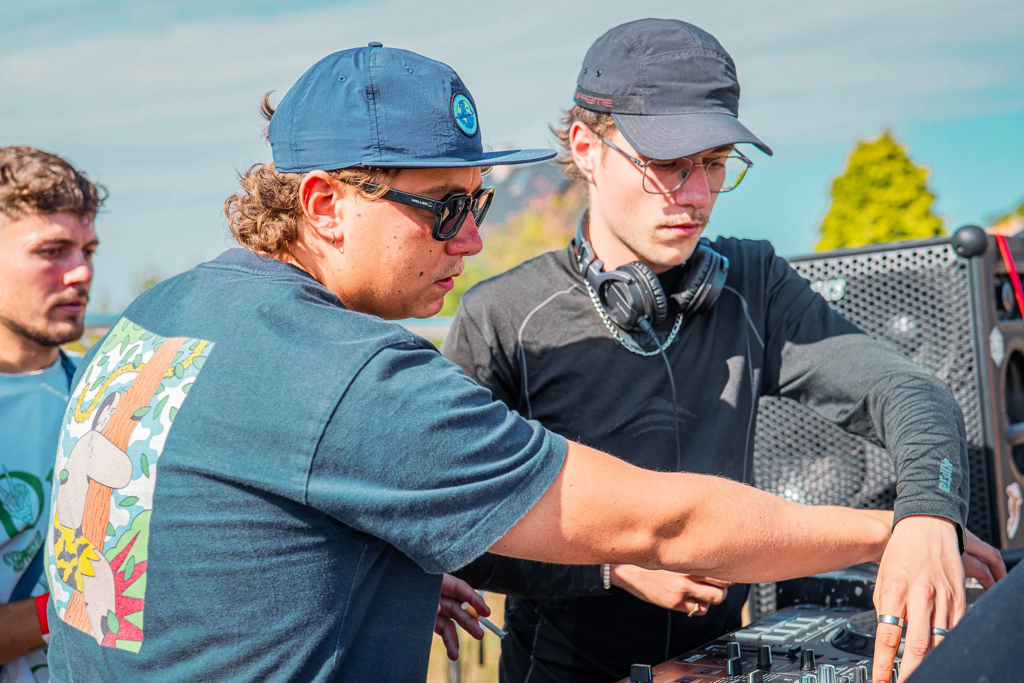 Trois jeunes hommes concentrés autour d'un équipement de DJ en extérieur, portant des casquettes et des écouteurs.