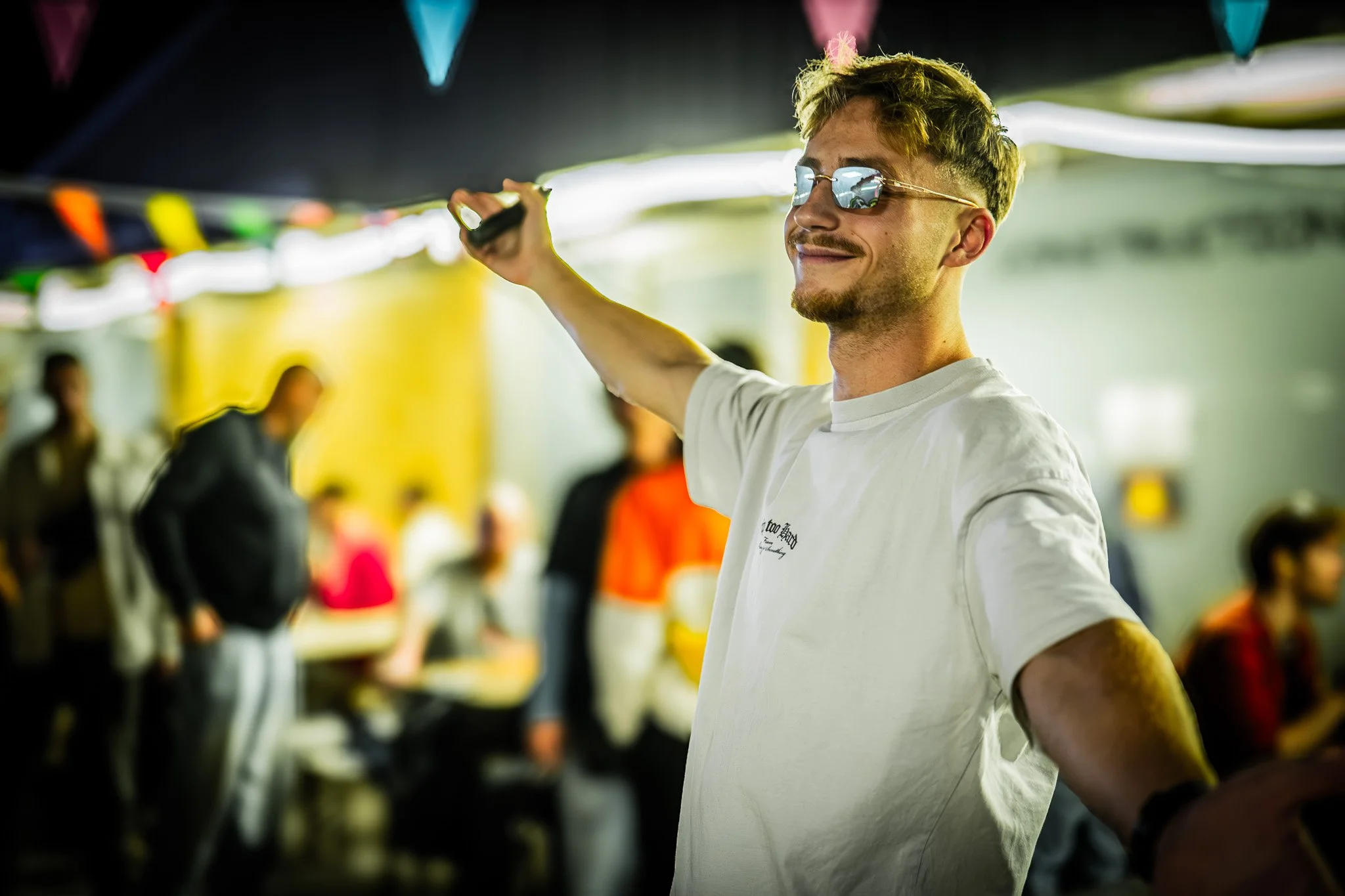 Un jeune homme portant des lunettes de soleil sourit dans une fête ou un rassemblement, avec des décorations colorées en arrière-plan et plusieurs personnes floues autour.