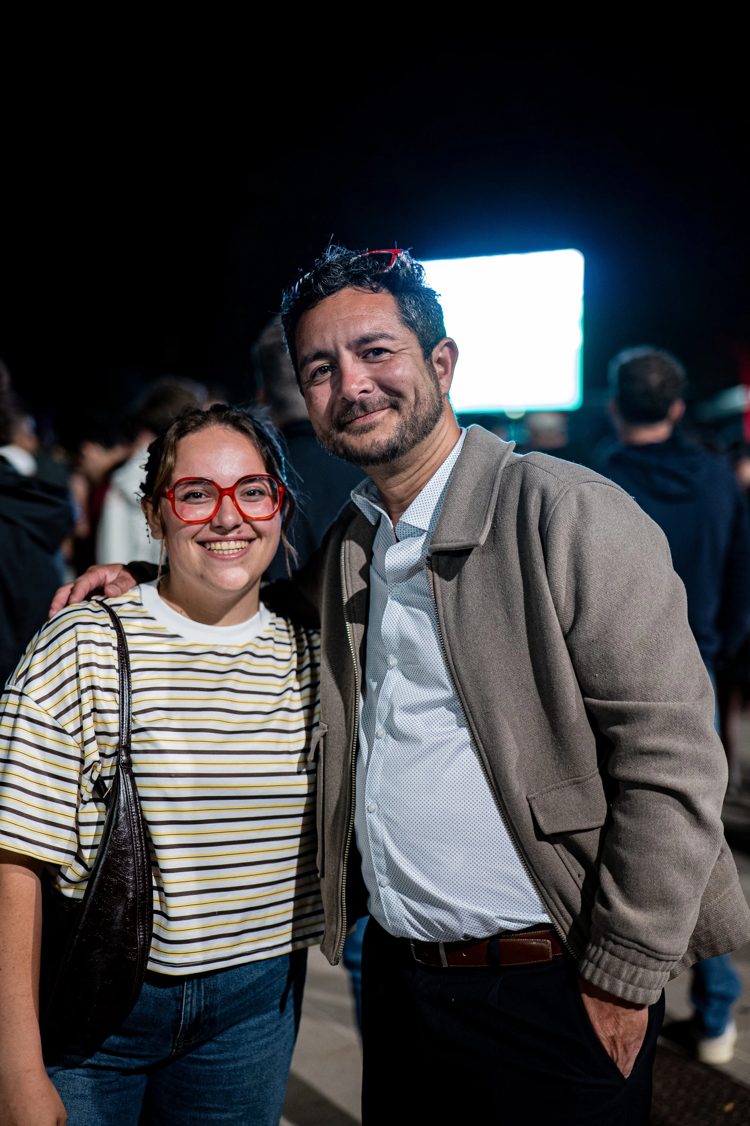 Deux personnes souriantes posent ensemble dans une ambiance sombre avec un écran lumineux en arrière-plan. L'une porte des lunettes rouges et un haut à rayures, l'autre un manteau beige et une chemise blanche. Ils semblent dans un lieu public ou une 