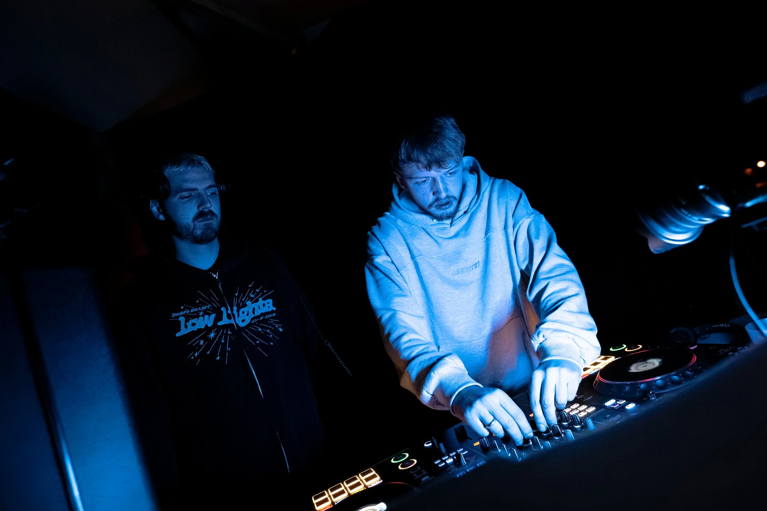 Deux hommes jouent de la musique en utilisant un équipement de DJ dans un environnement sombre, éclairé par des lumières bleues et colorées.