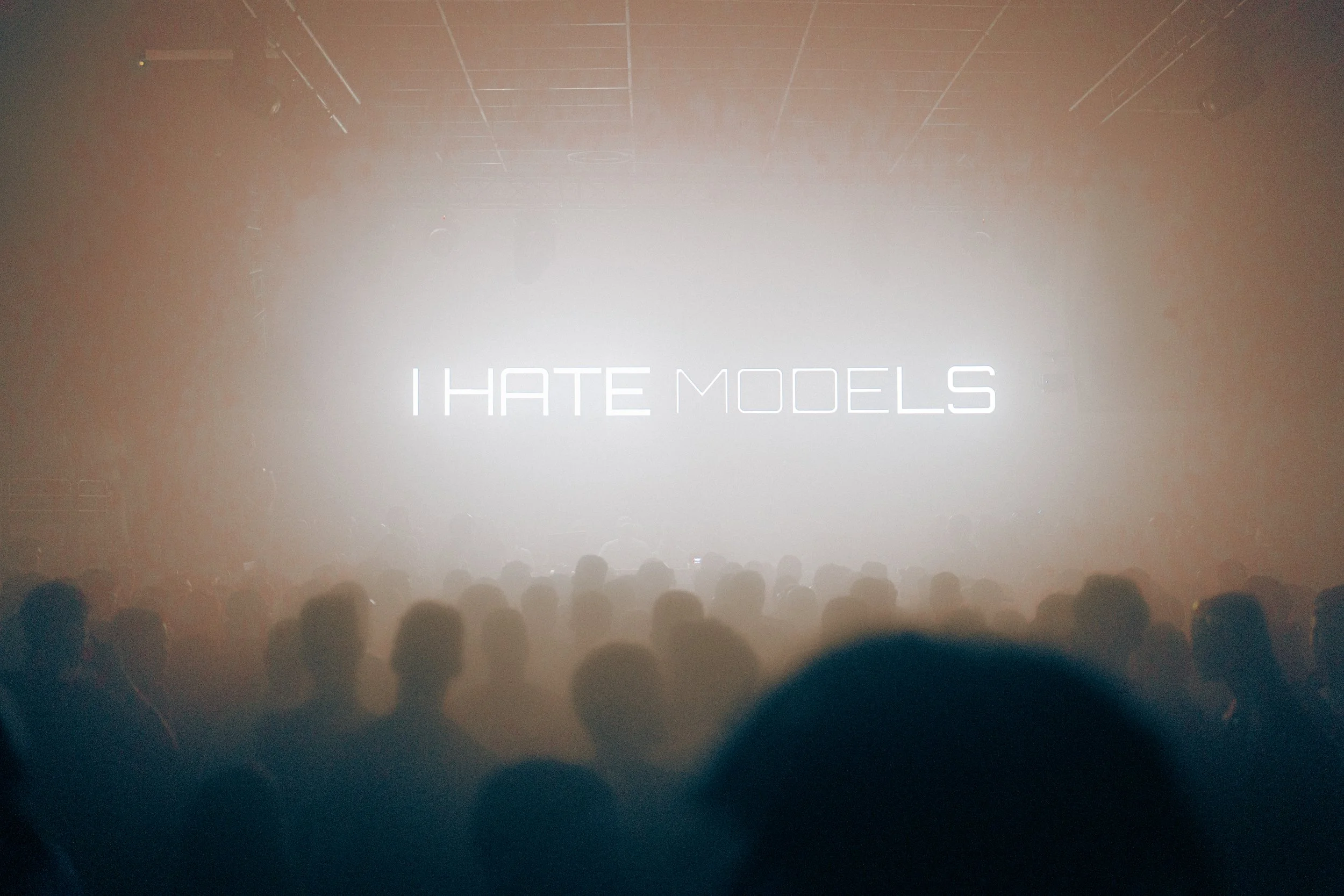 Une scène d'un concert ou d'un spectacle avec une audience vue de dos et un écran lumineux affichant le texte "I HATE MODELS".