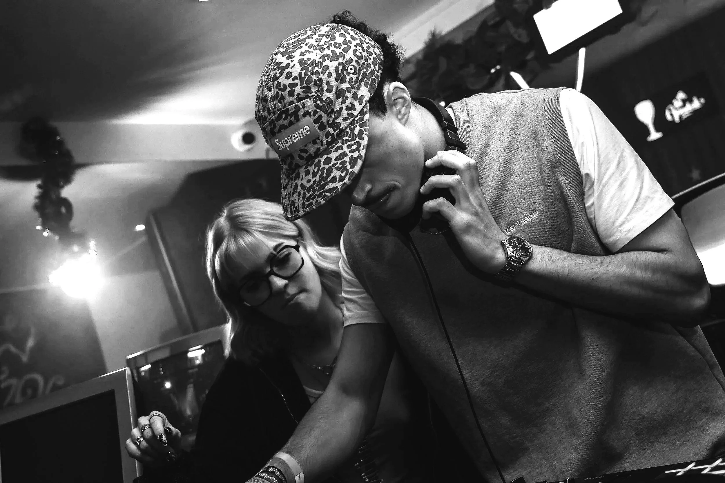 Deux personnes, une femme avec des lunettes et une autre personne portant une casquette à motifs léopard avec le logo Supreme, assises l'une à côté de l'autre, dans un cadre intérieur.