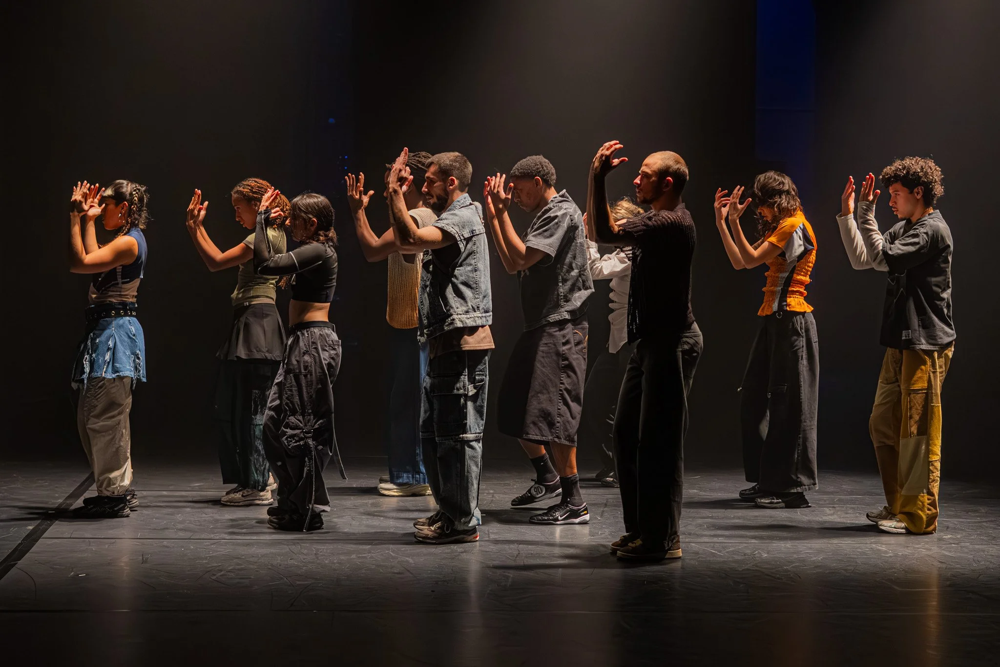 Groupe de personnes debout en ligne sur scène, posant avec les mains levées, dans une scène sombre, peut-être pour une performance ou une répétition.